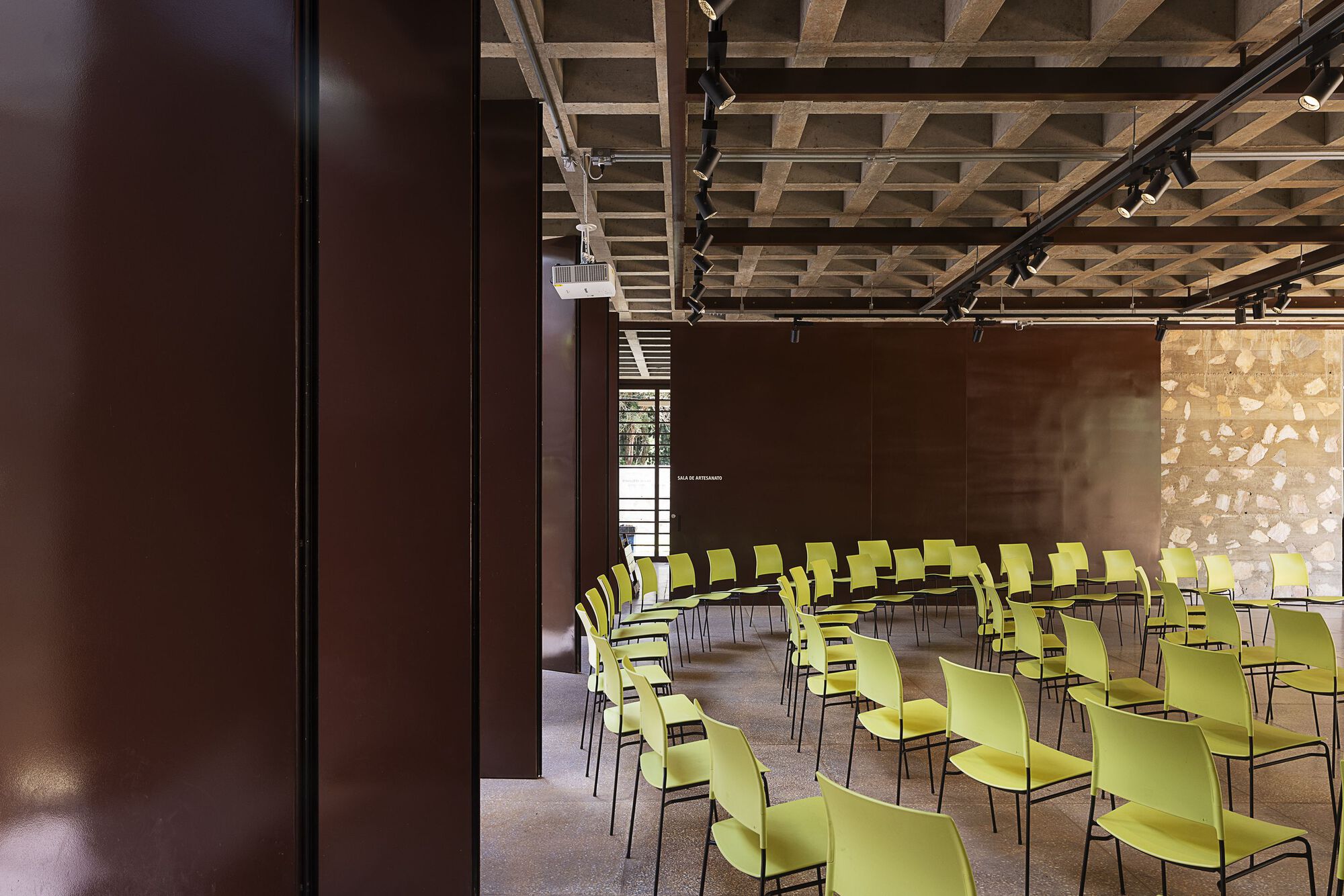 Curved rows of green chairs in a hall with rust-toned steel panels, a rubble stone wall and a gridded concrete ceiling