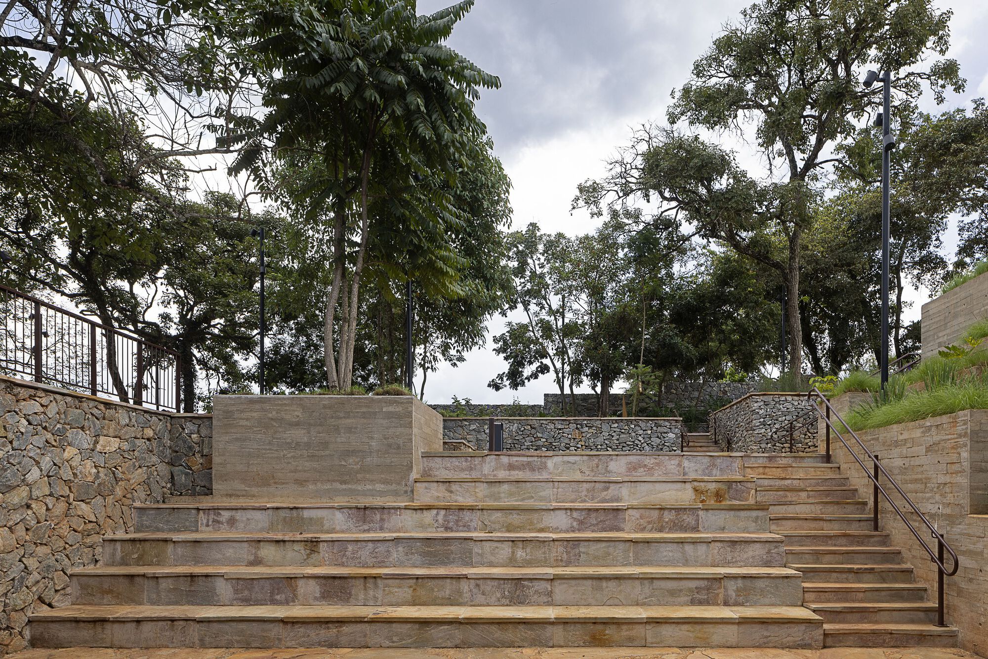 Wide stone steps with planter blocks, rubble retaining walls and a metal handrail beneath tall trees and an overcast sky