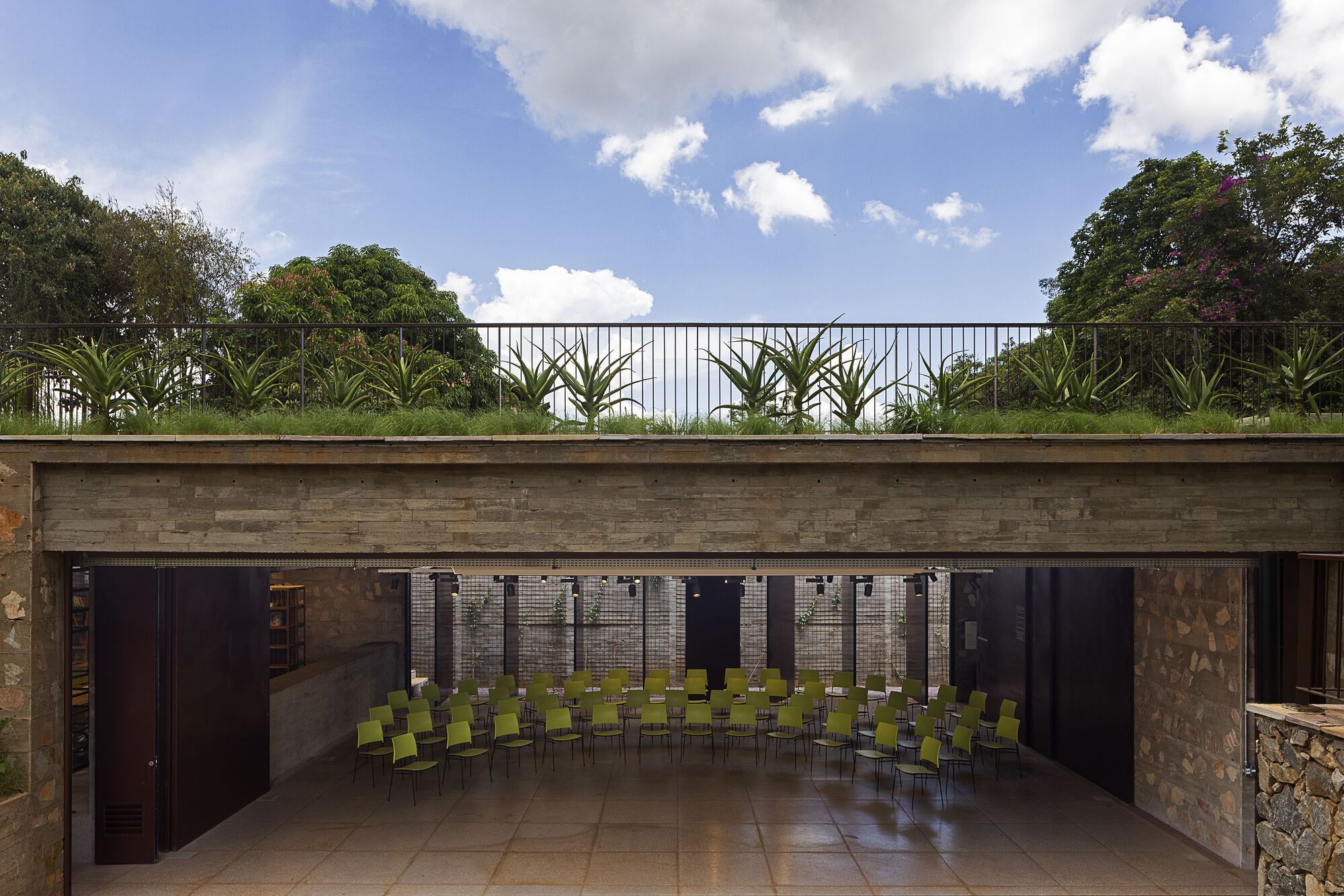 Open-sided hall with rows of green chairs beneath a stone-faced beam and a green roof planted with agave under blue sky
