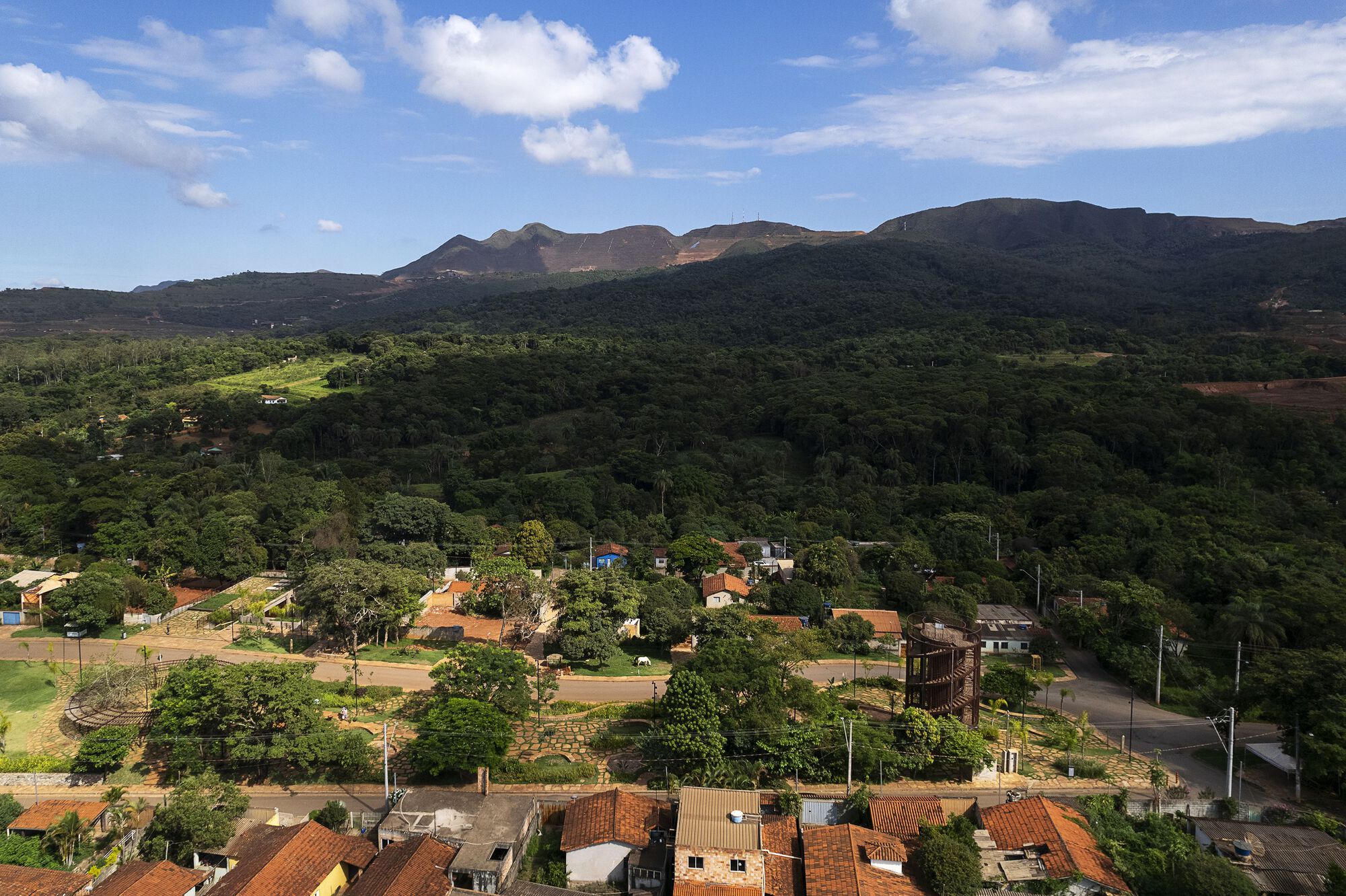 Aerial view of a hillside town with terracotta roofs, a timber lattice tower among trees and forested mountains under scattered clouds