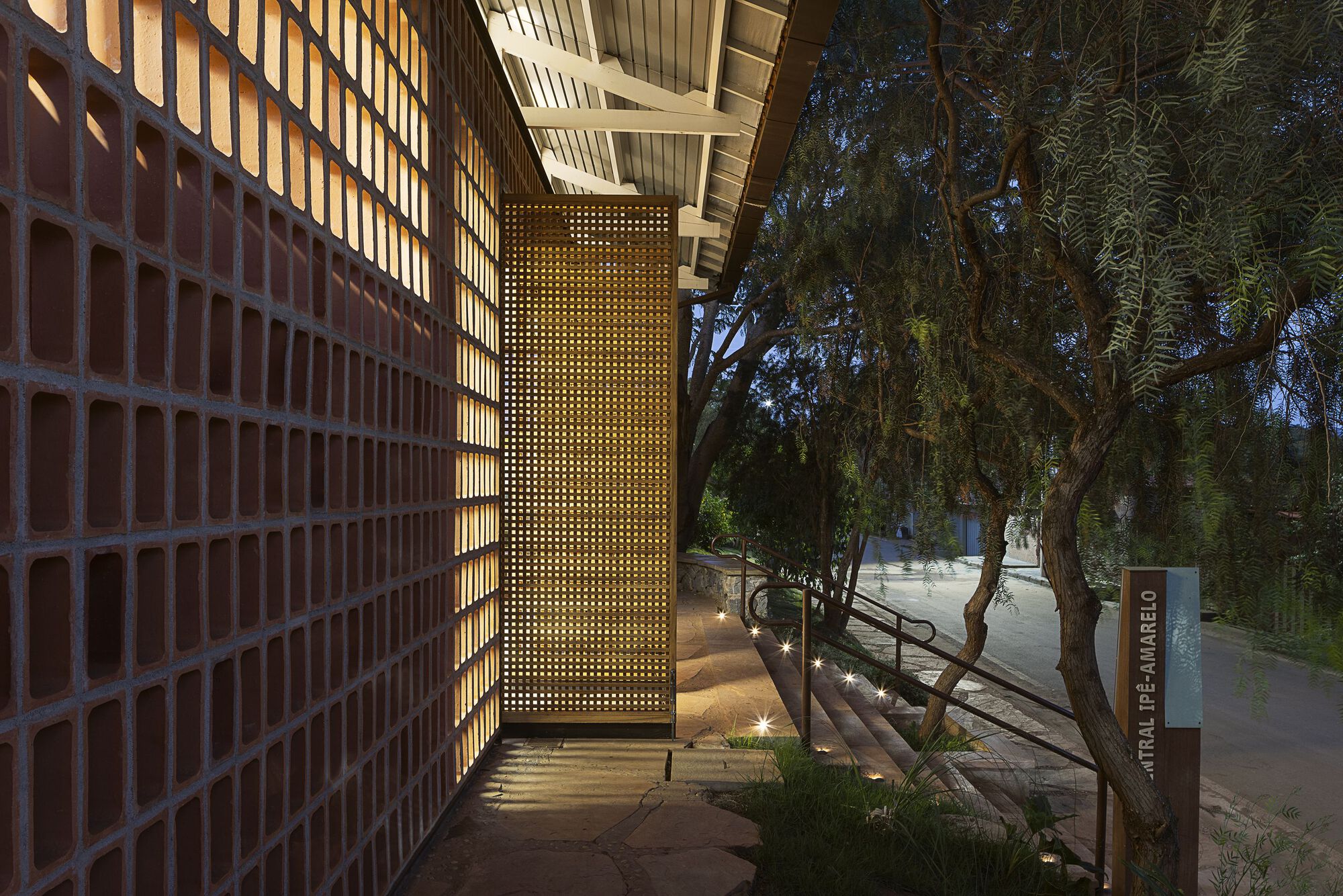 Perforated red clay block wall glowing from within beside a timber lattice screen and a stepped path at dusk