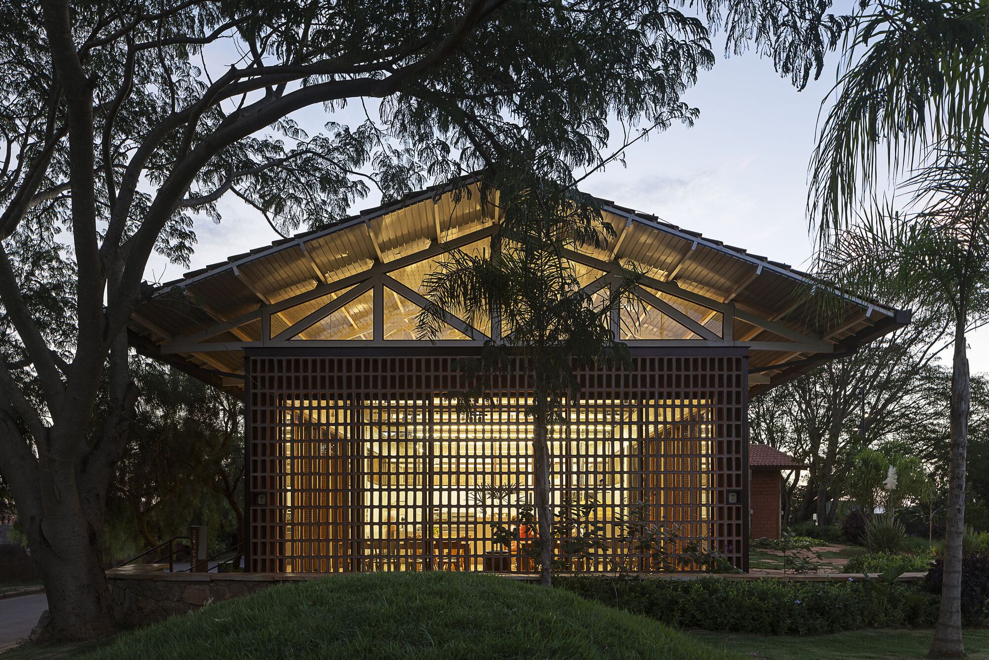 Gabled pavilion with a timber lattice facade and exposed trusses lit from inside, framed by large trees at twilight