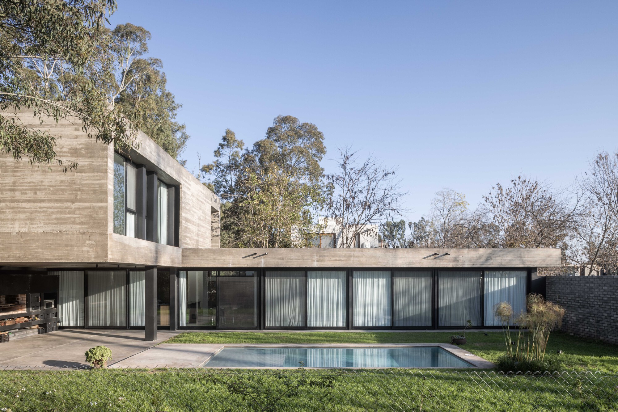 Garden facade of board-formed concrete volume over a glazed ground floor with white curtains, a rectangular pool in the foreground lawn under clear sky