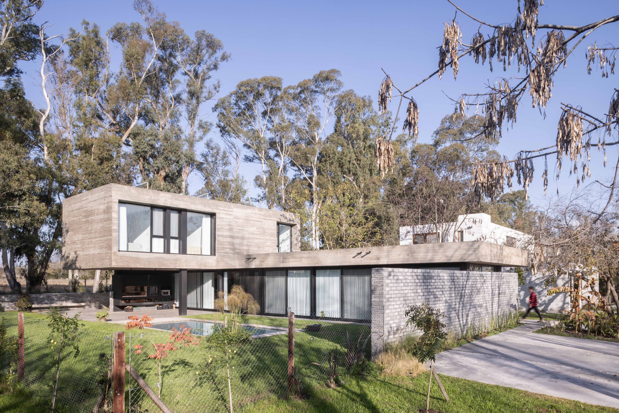 Corner view of the concrete upper volume and grey brick wall, seen through wire fencing and eucalyptus trees as a person walks past