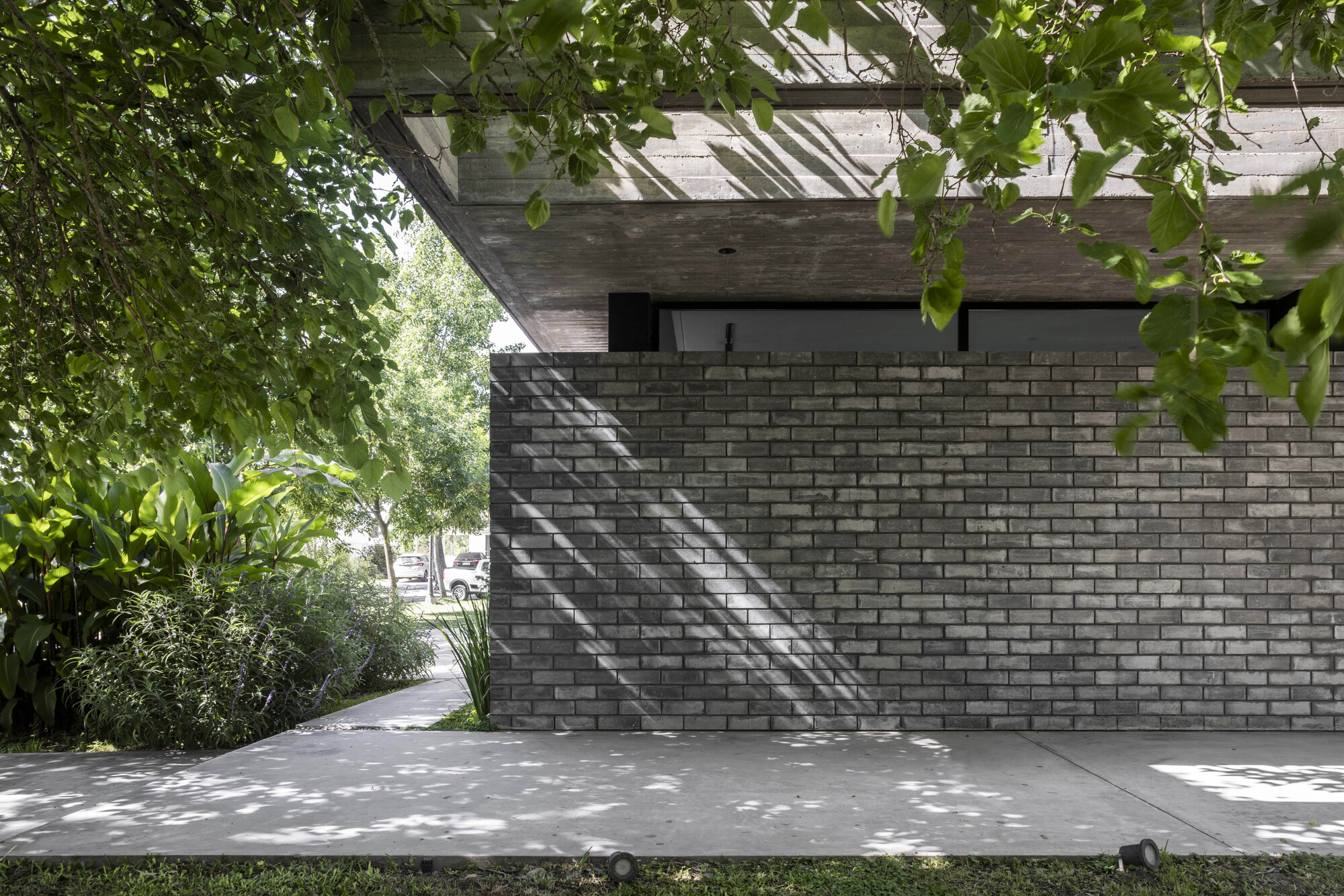 Grey brick wall beneath a concrete slab overhang, with dappled leaf shadows falling across the wall and the paved path