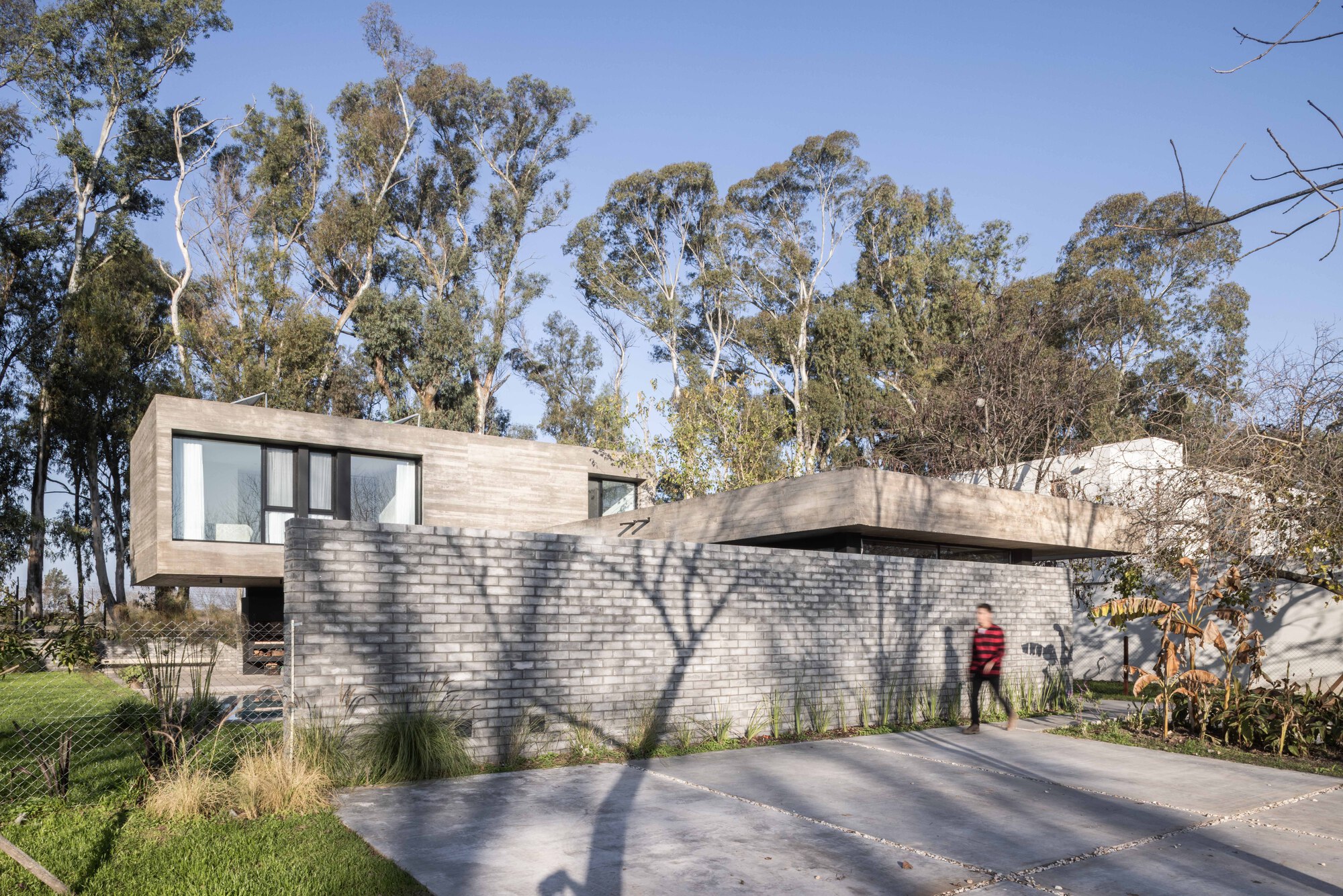 Street side of the grey brick wall and concrete upper volume, with a person in a red sweater walking past in tree shadows