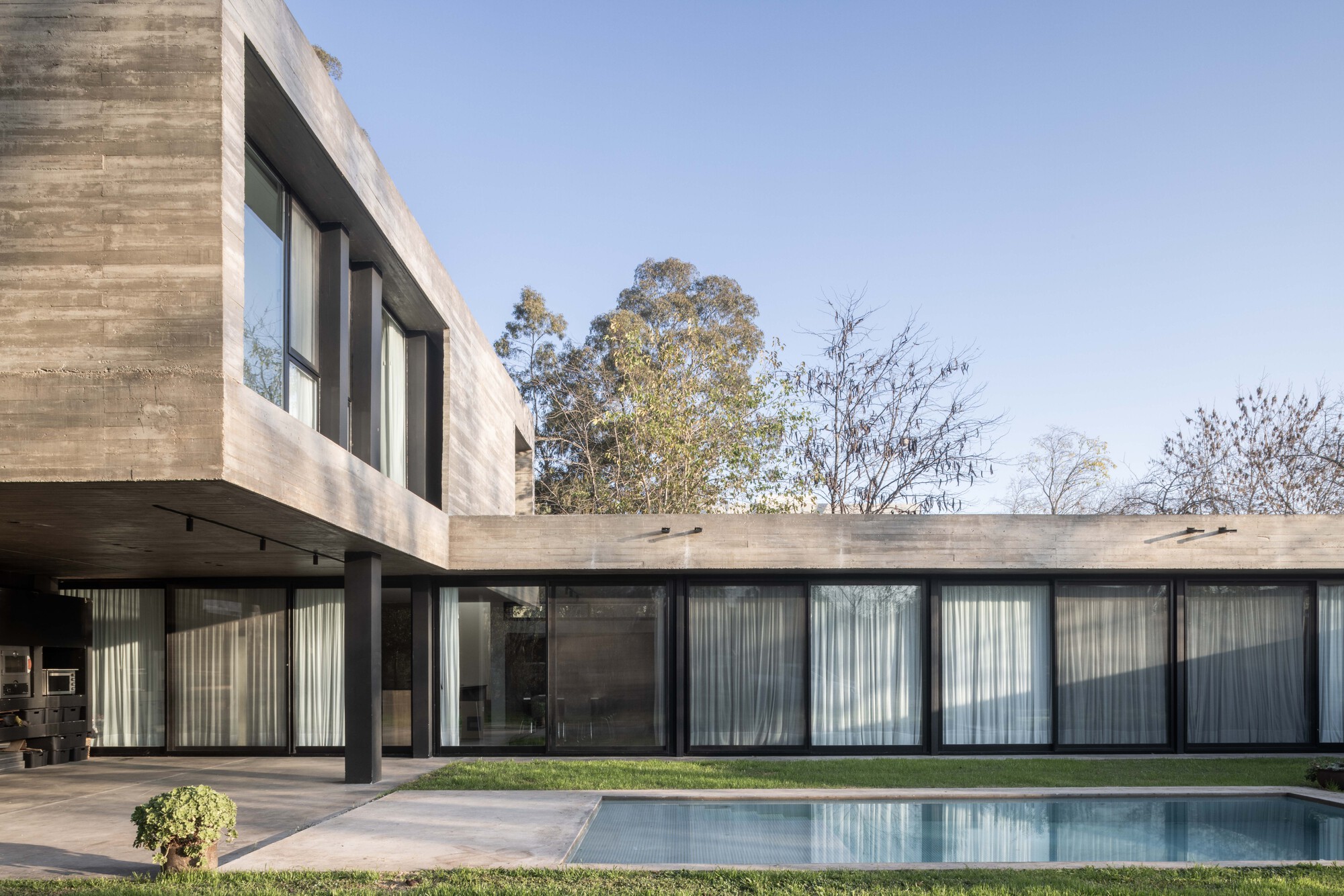Cantilevered board-formed concrete upper volume above a glazed ground floor with sheer curtains, beside a pool under a clear blue sky