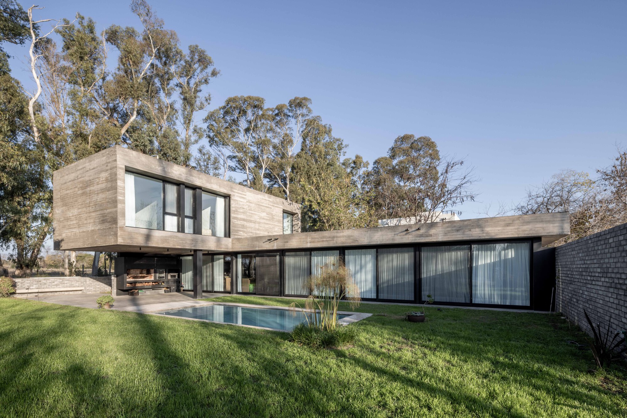 L-shaped concrete house seen across a lawn, with a pool at its corner and tall eucalyptus trees behind