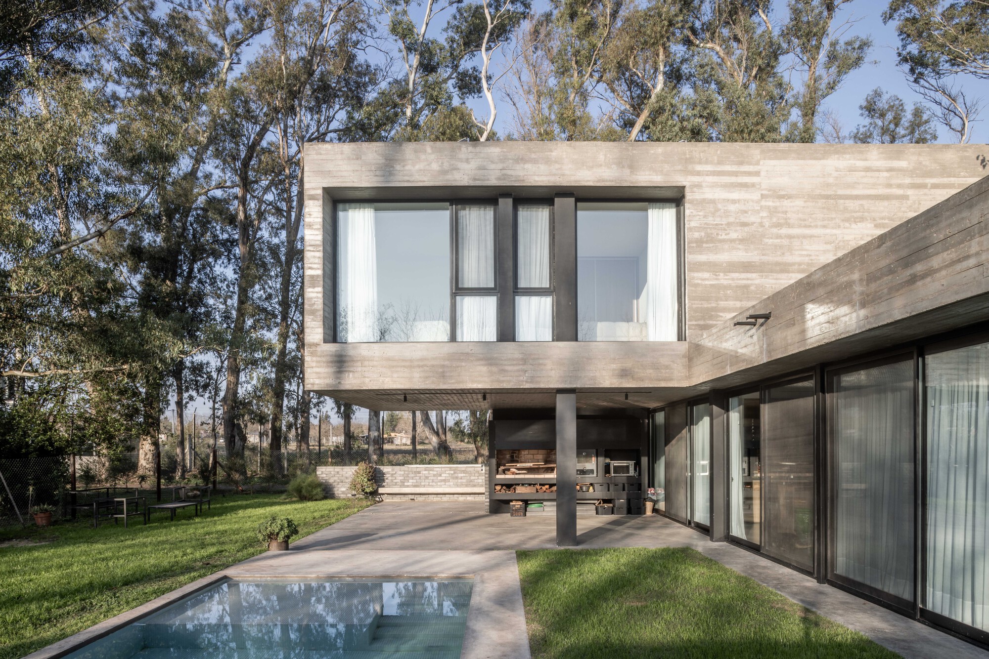 Concrete upper volume on a steel column above a covered terrace with a built-in grill and firewood storage, pool in the foreground