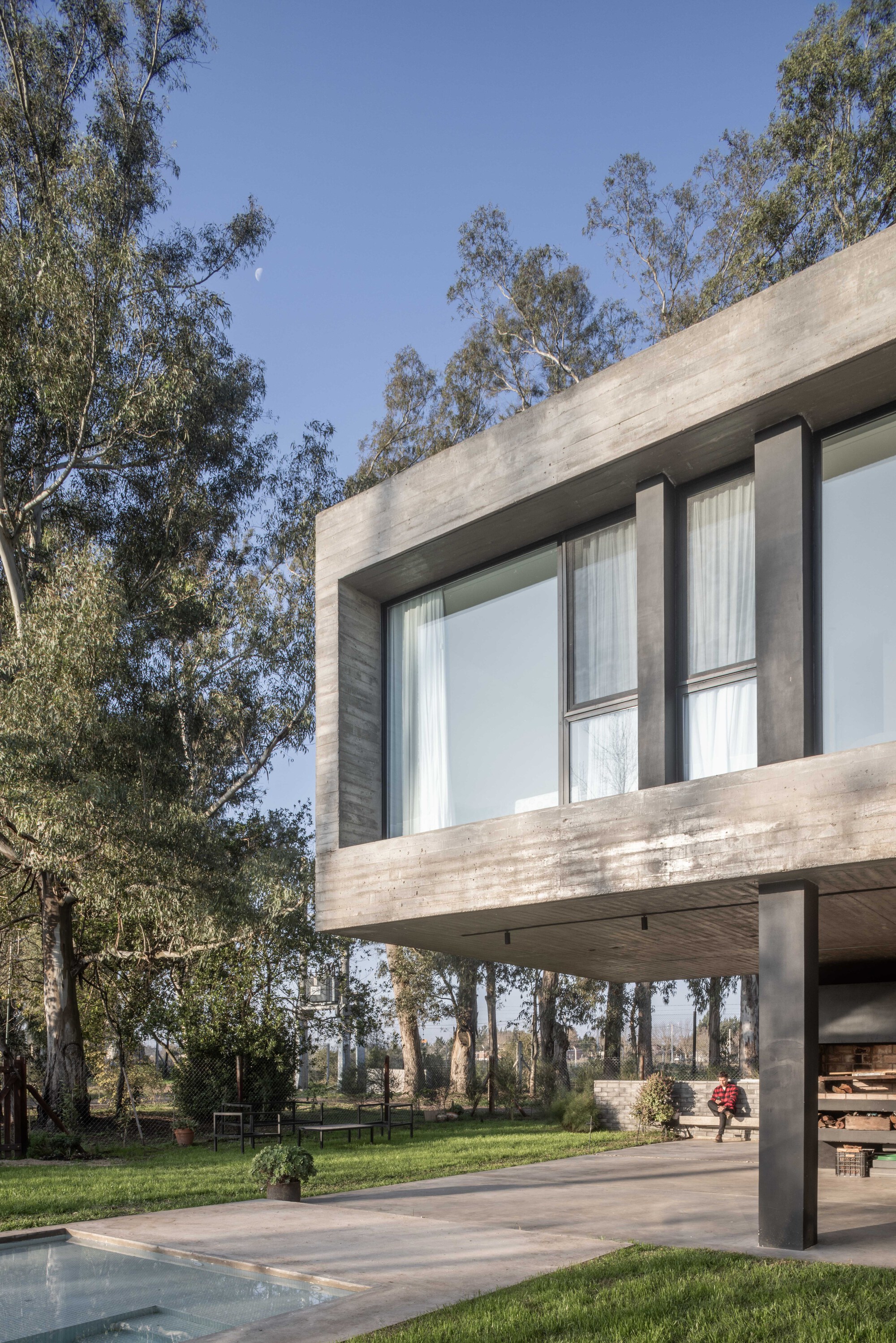Low-angle view of the board-formed concrete box and dark steel columns, with a person seated on a bench under eucalyptus trees