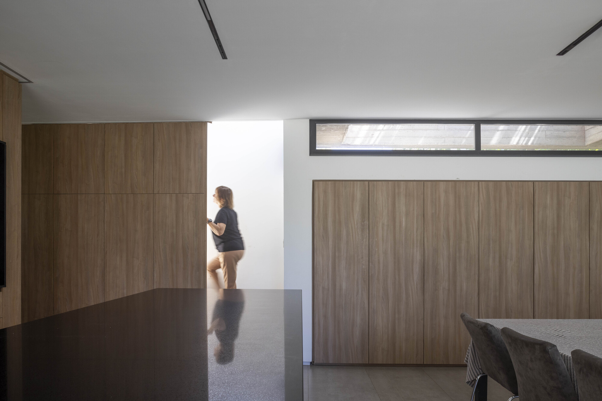 Kitchen island in dark stone beside full-height timber cabinet doors, with a woman walking toward a bright stairwell opening