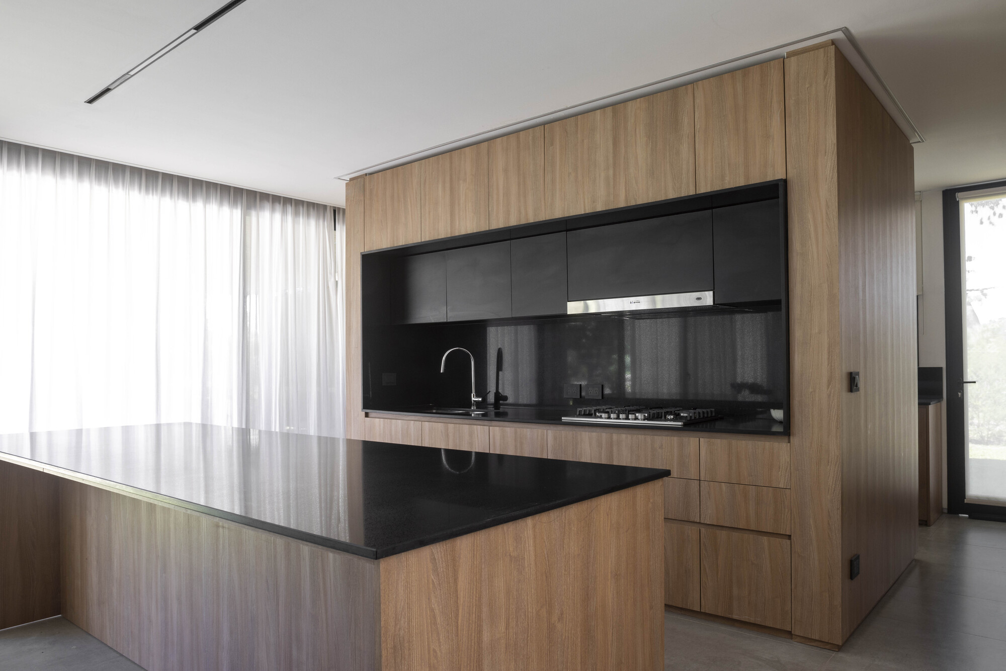 Timber-clad kitchen block with black stone island and backsplash, beside sheer curtains diffusing daylight