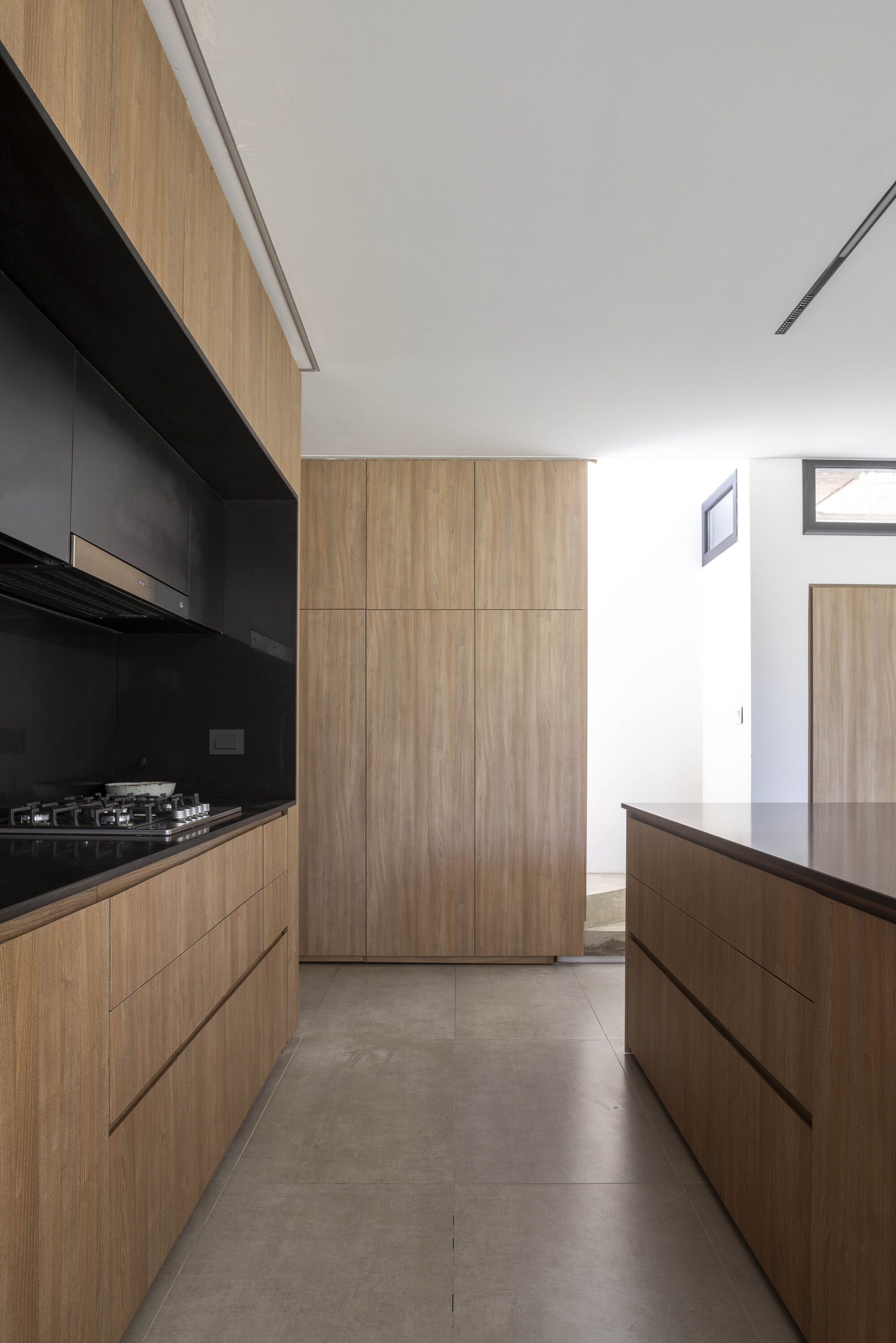 Narrow kitchen aisle between timber cabinets with a black cooktop niche and tall storage wall, lit from a side window
