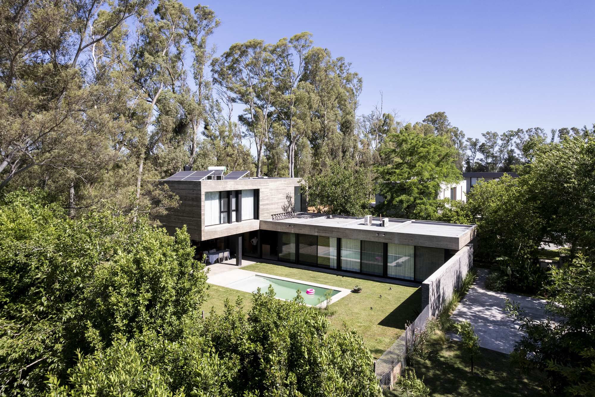 Garden elevation of the upper volume cantilevered over a glazed ground floor, with a lawn and turquoise pool in midday sun