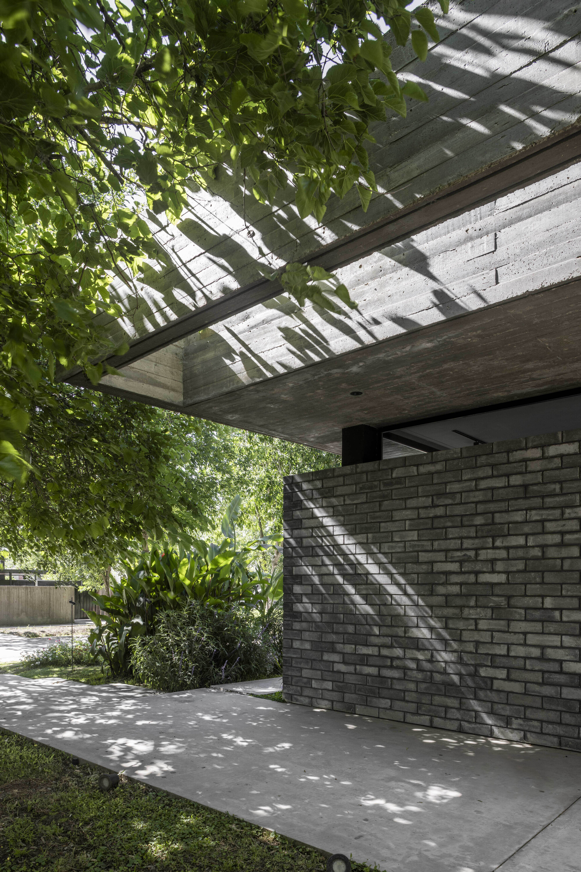 Board-formed concrete roof overhang above a gray brick wall, with tree shadows scattered across the concrete path