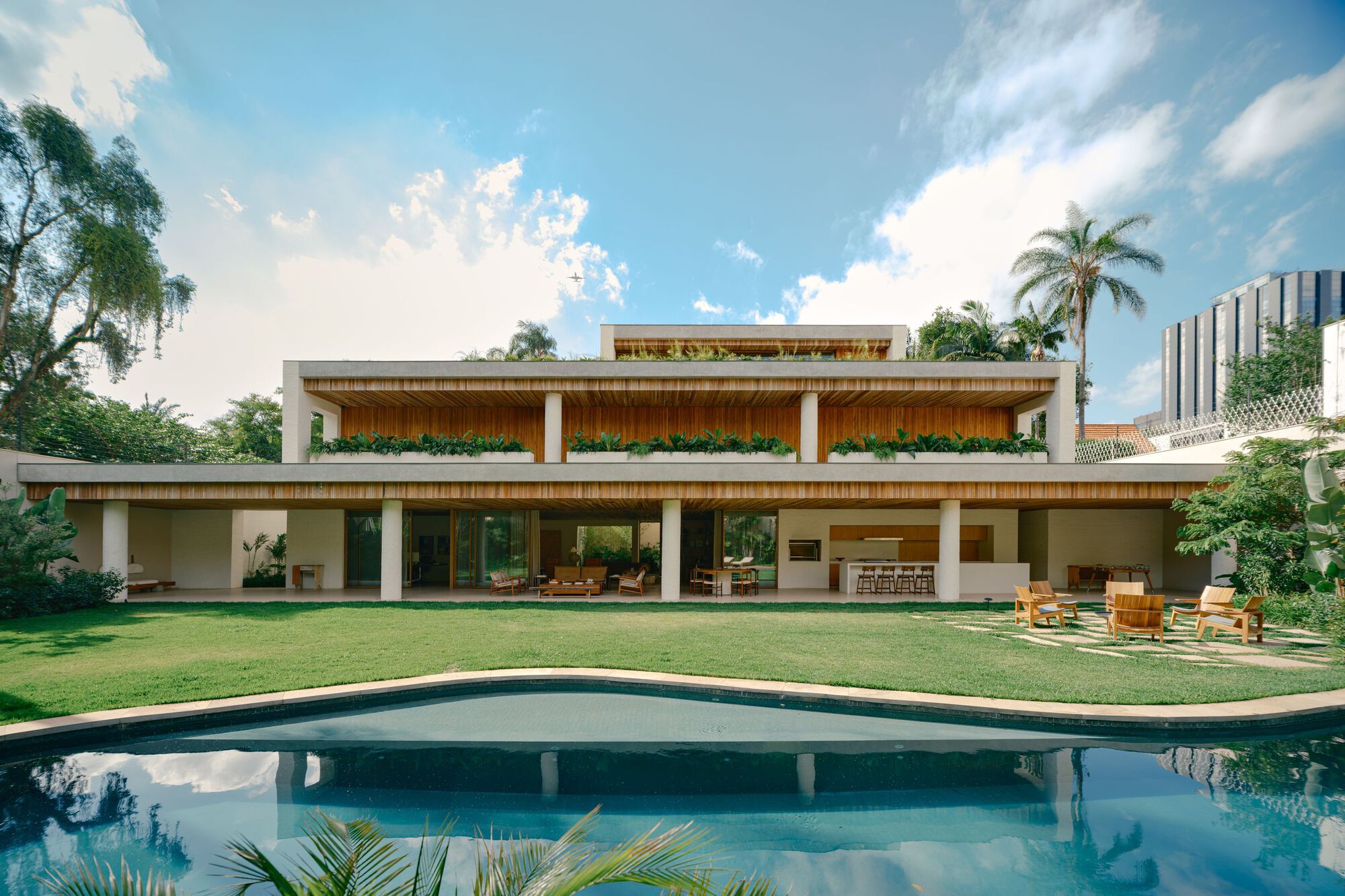 Rear facade with timber-lined soffits and planter boxes above a lawn and curved pool under a bright cloudy sky