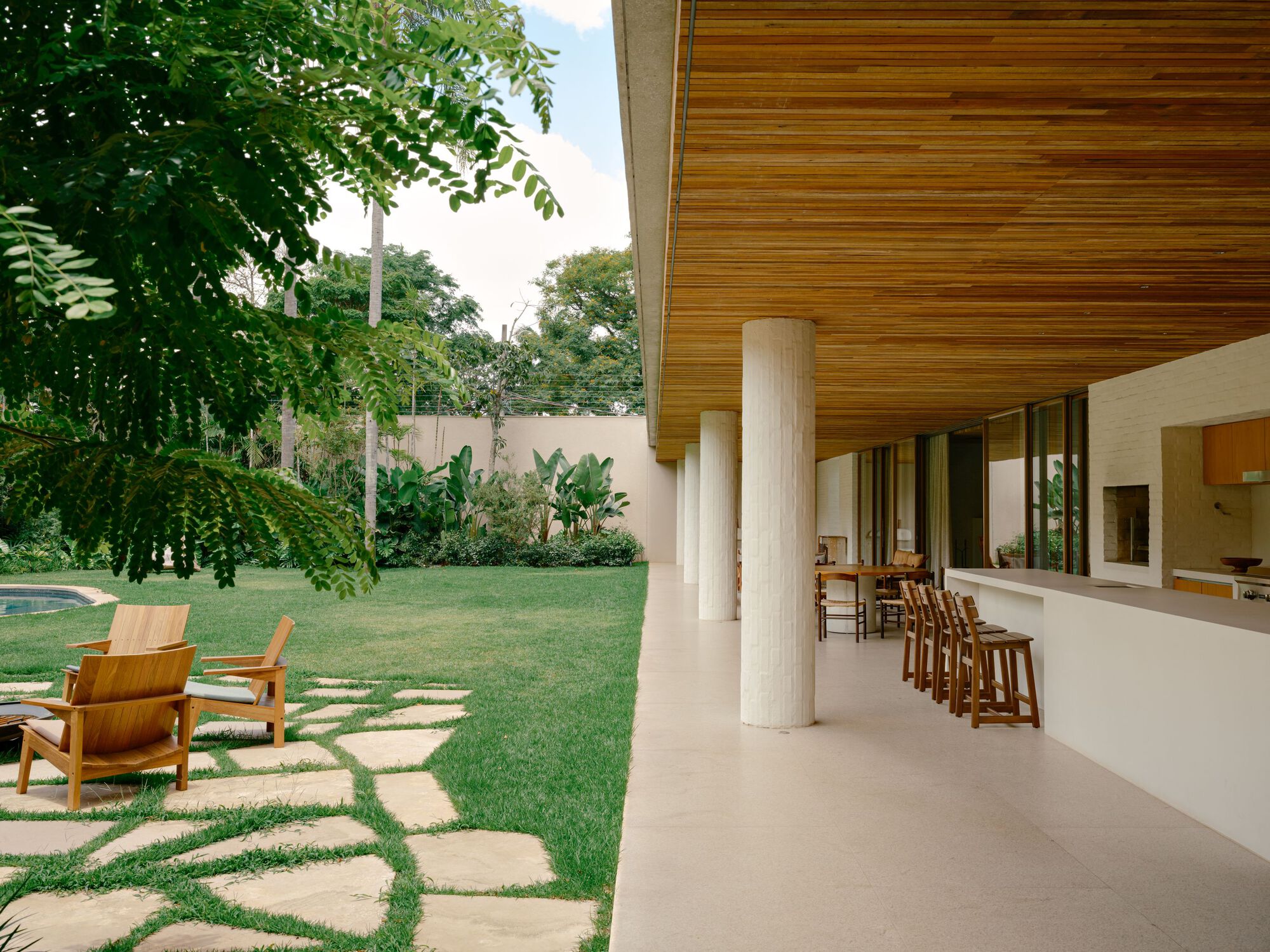 Covered terrace with round white brick columns, timber ceiling and bar stools beside a lawn with stone stepping pads