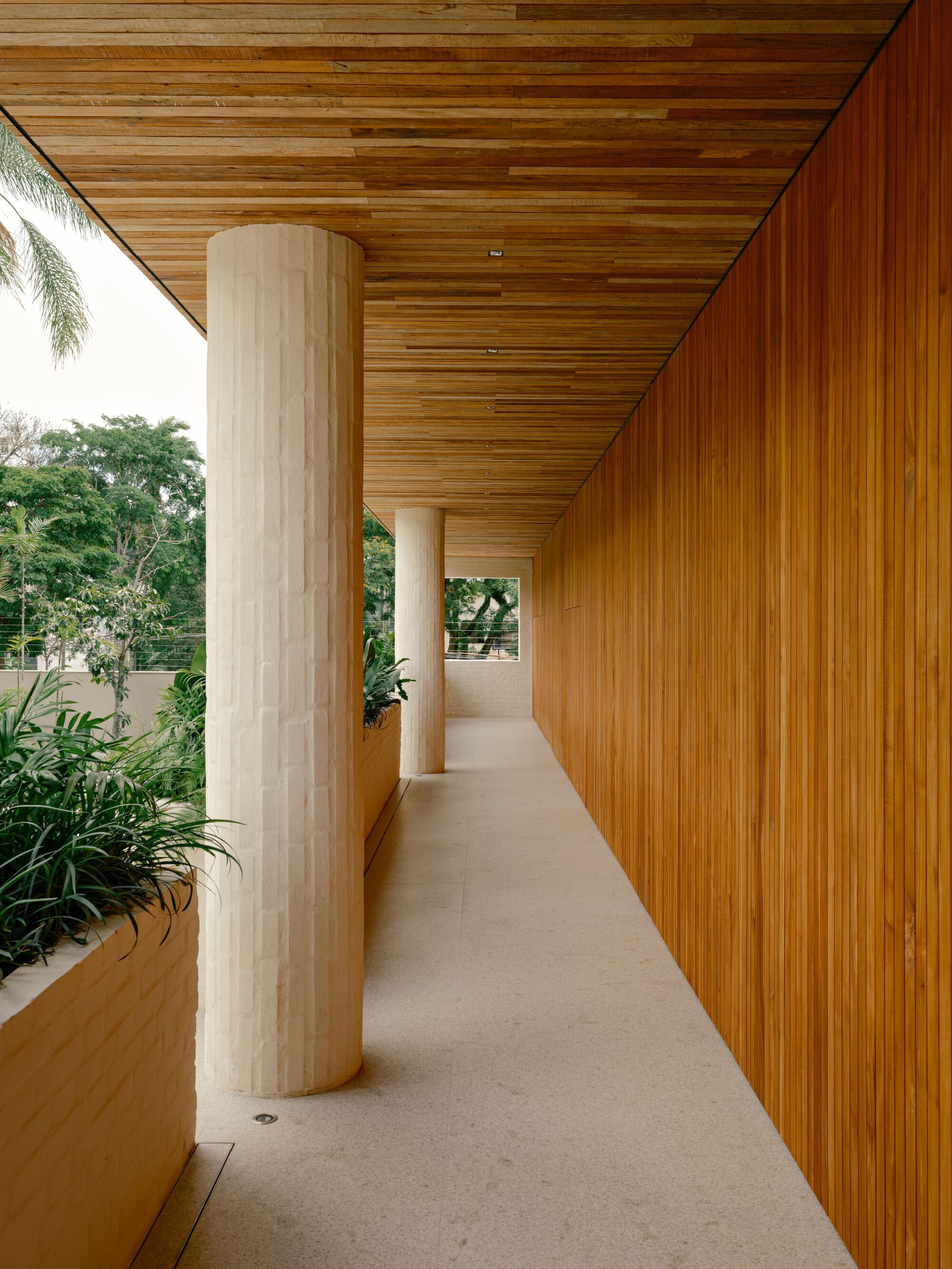 Upper walkway lined with round brick columns, planters and vertical timber cladding, with treetops visible beyond in daylight
