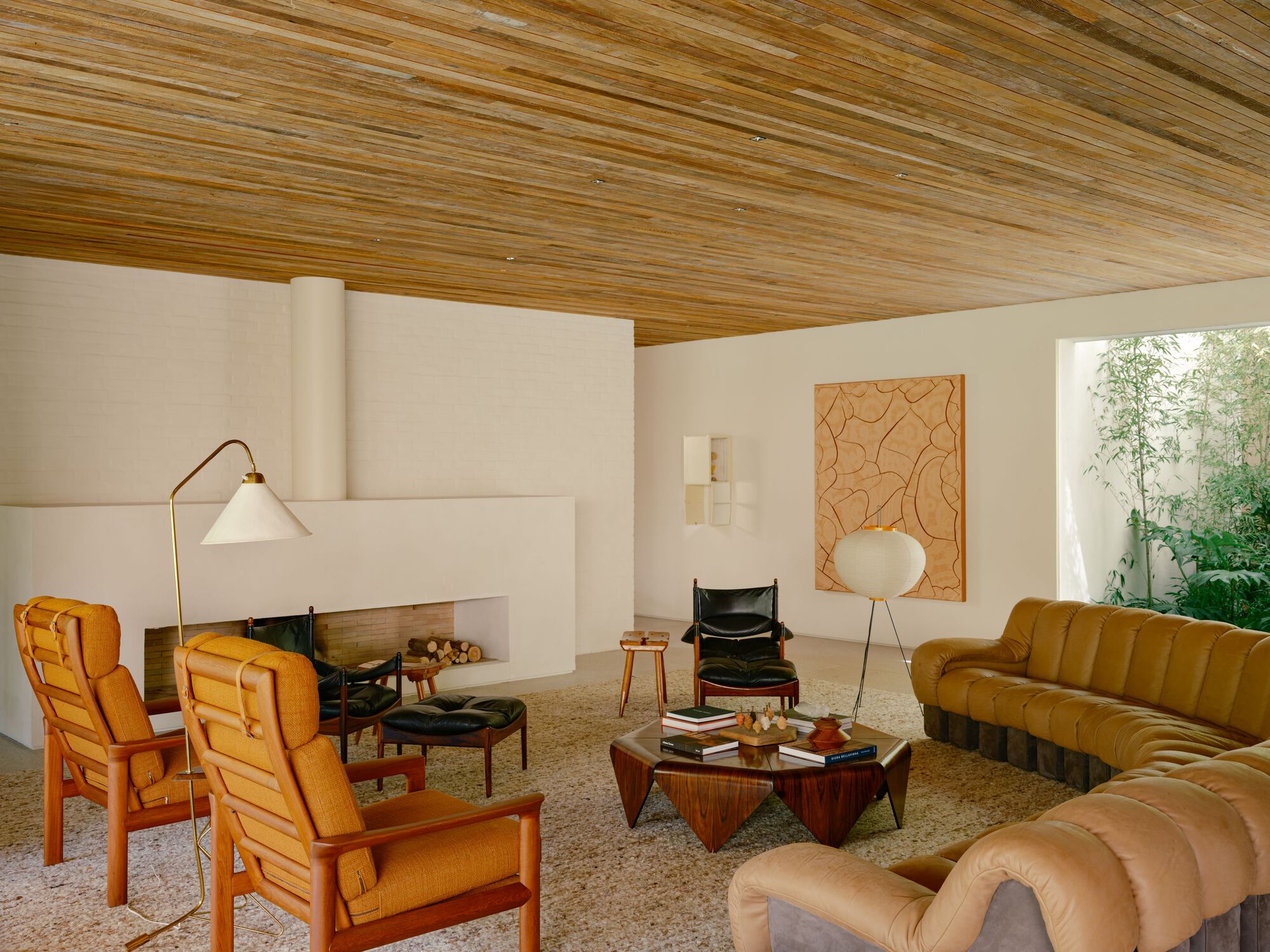 Living room with a white hearth, orange wood-framed armchairs, a low wooden table and a timber ceiling in soft daylight