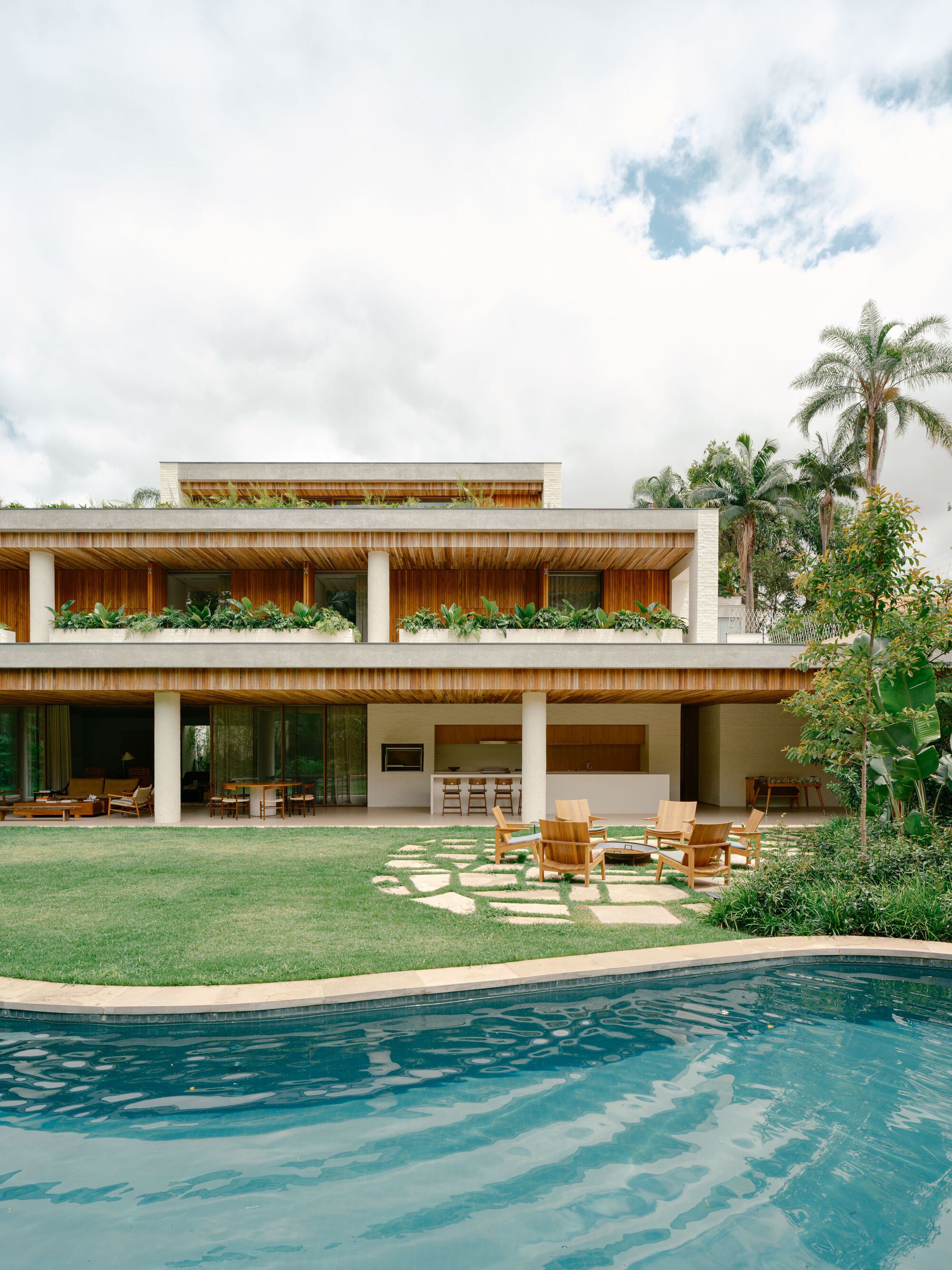 Rear facade of stacked timber-slatted floors with planter boxes, stepping stones and a curved pool edge under a cloudy sky
