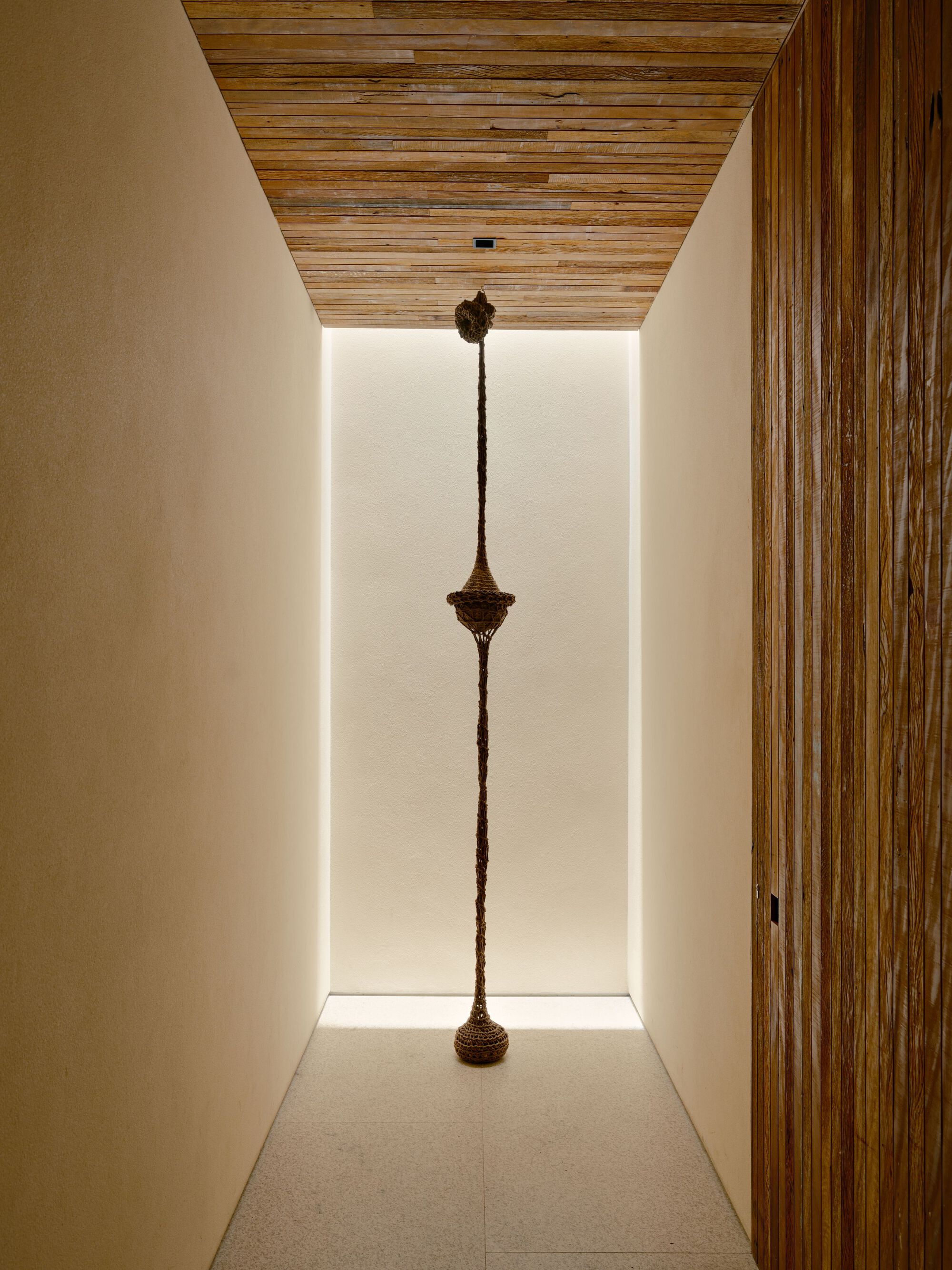 Narrow corridor with a reclaimed wood ceiling and a hanging woven sculpture lit by a glowing white wall slot