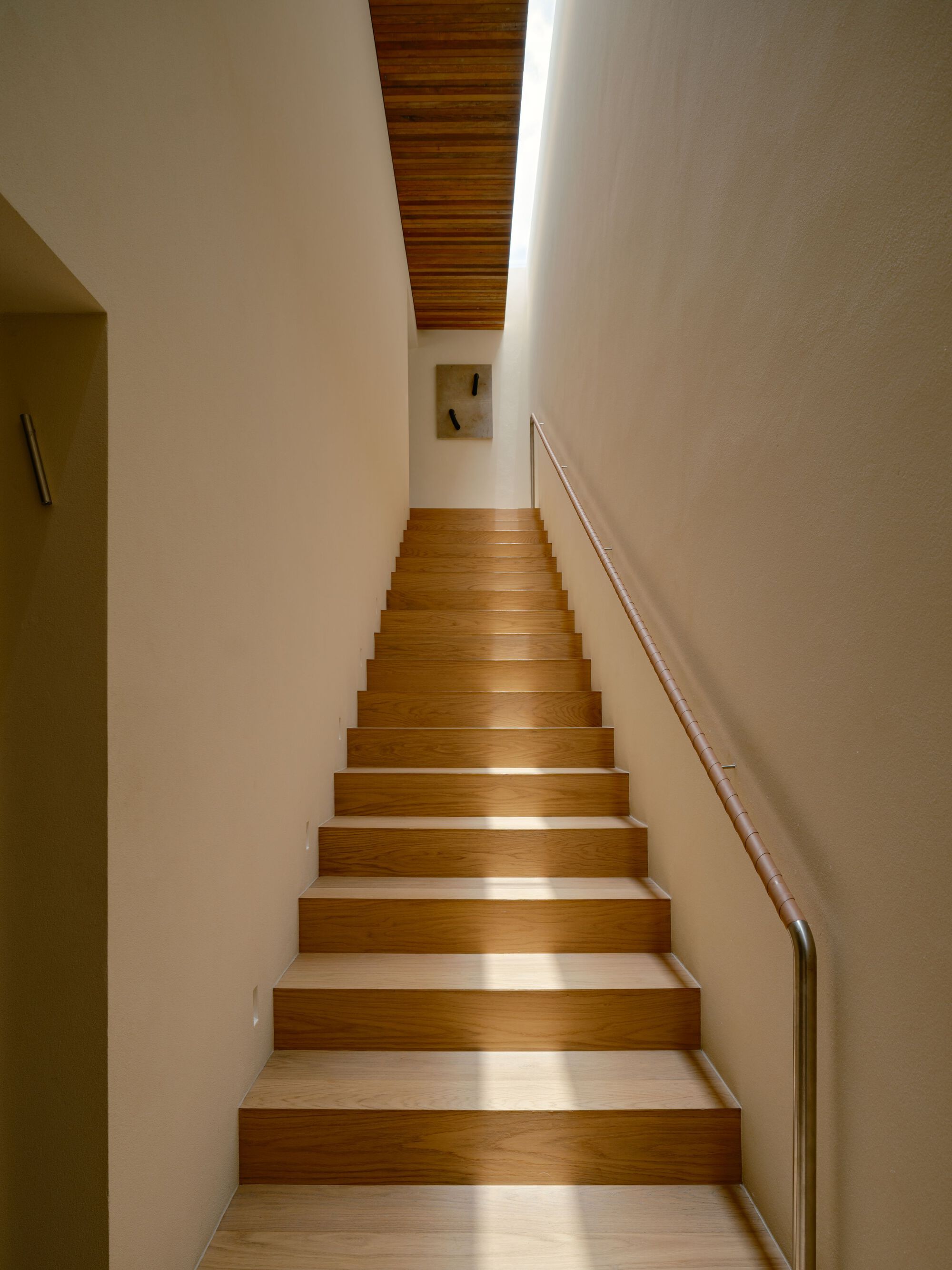 Straight oak staircase with a leather-wrapped steel handrail rising toward a skylit slot and a small artwork on the wall