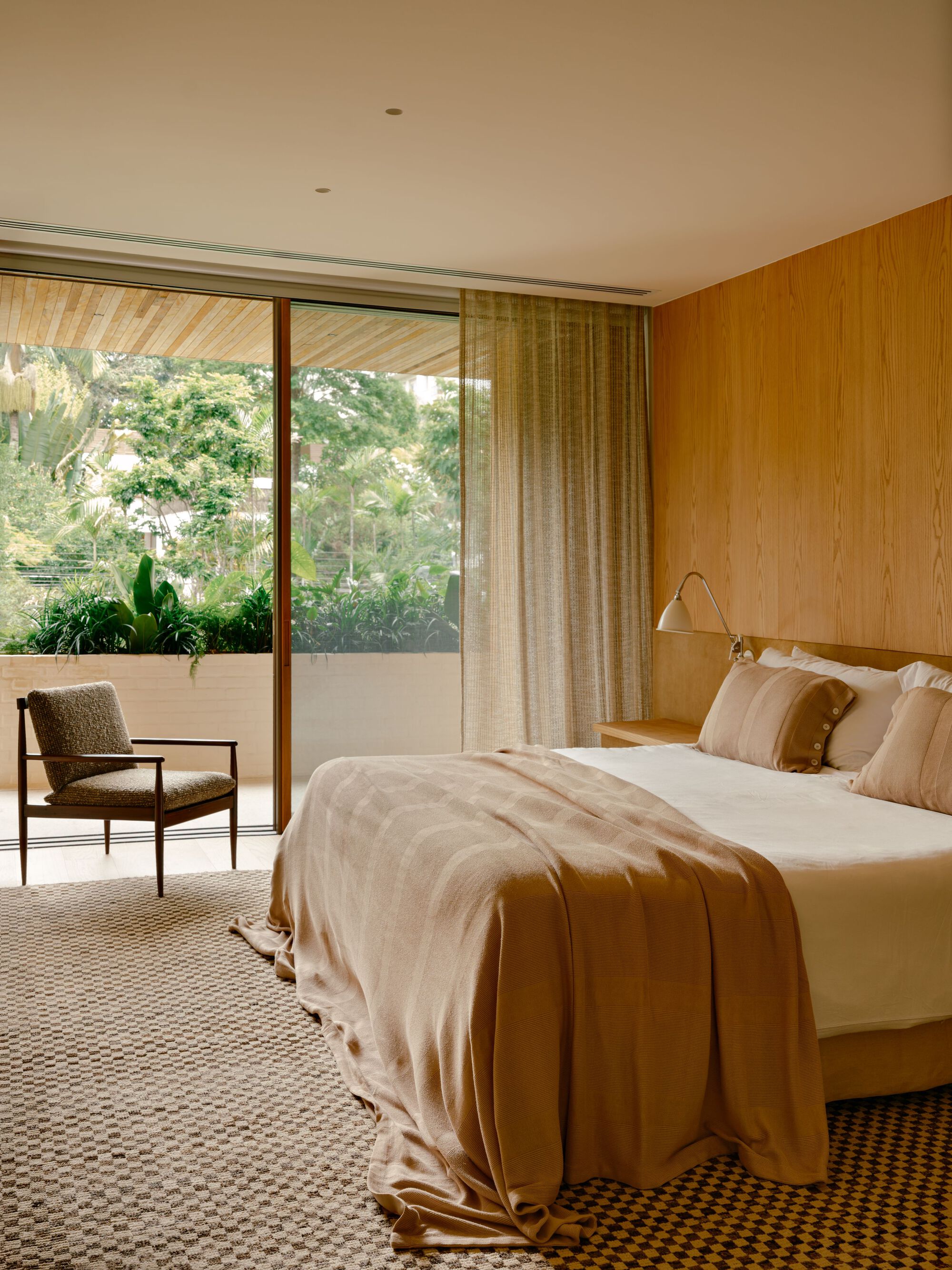 Bedroom with oak-paneled wall, checked rug and sheer curtain beside a sliding glass door opening to green foliage
