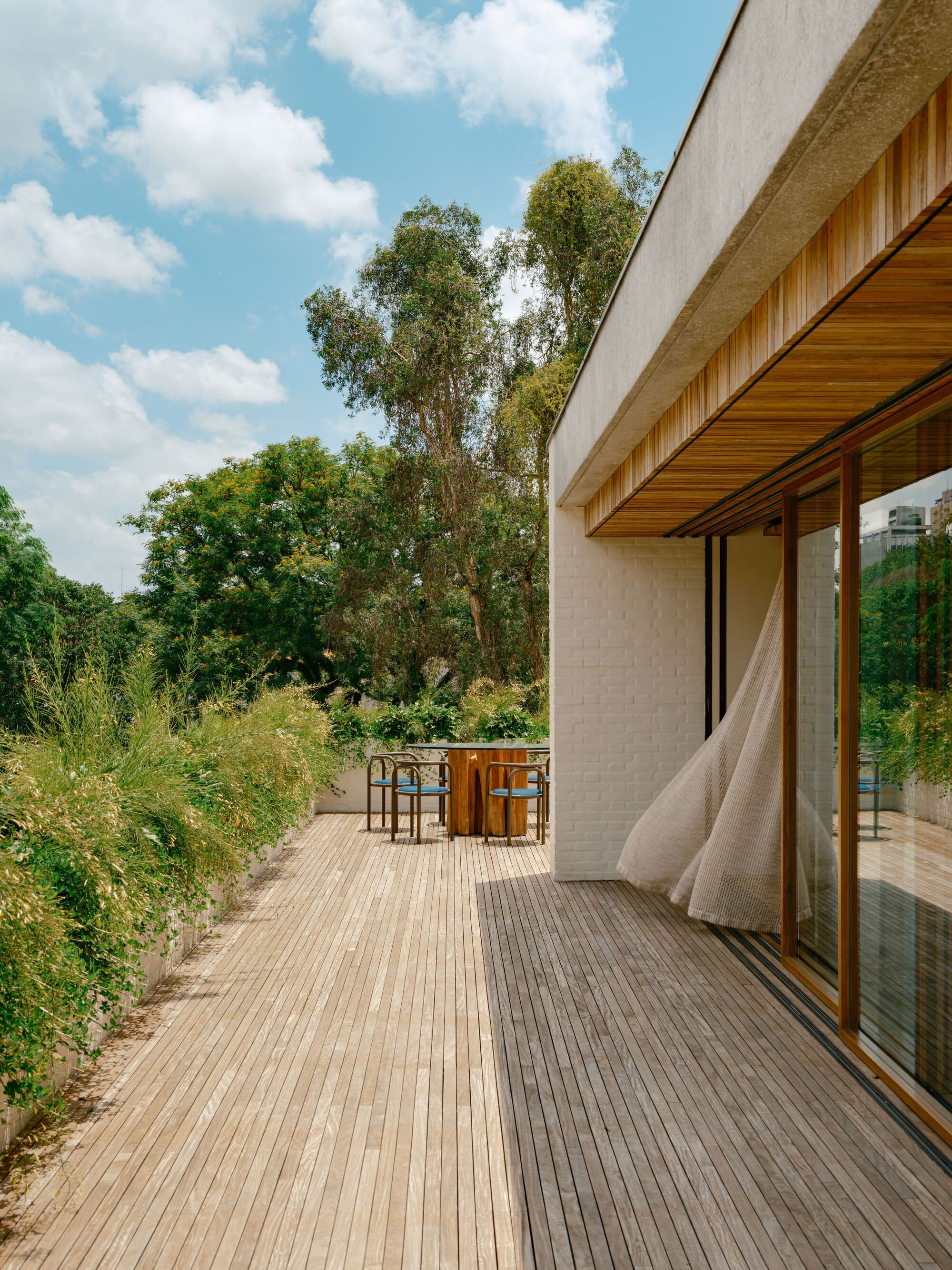 Slatted timber deck along a white brick wall with a billowing curtain, dining table and tall trees in afternoon sun