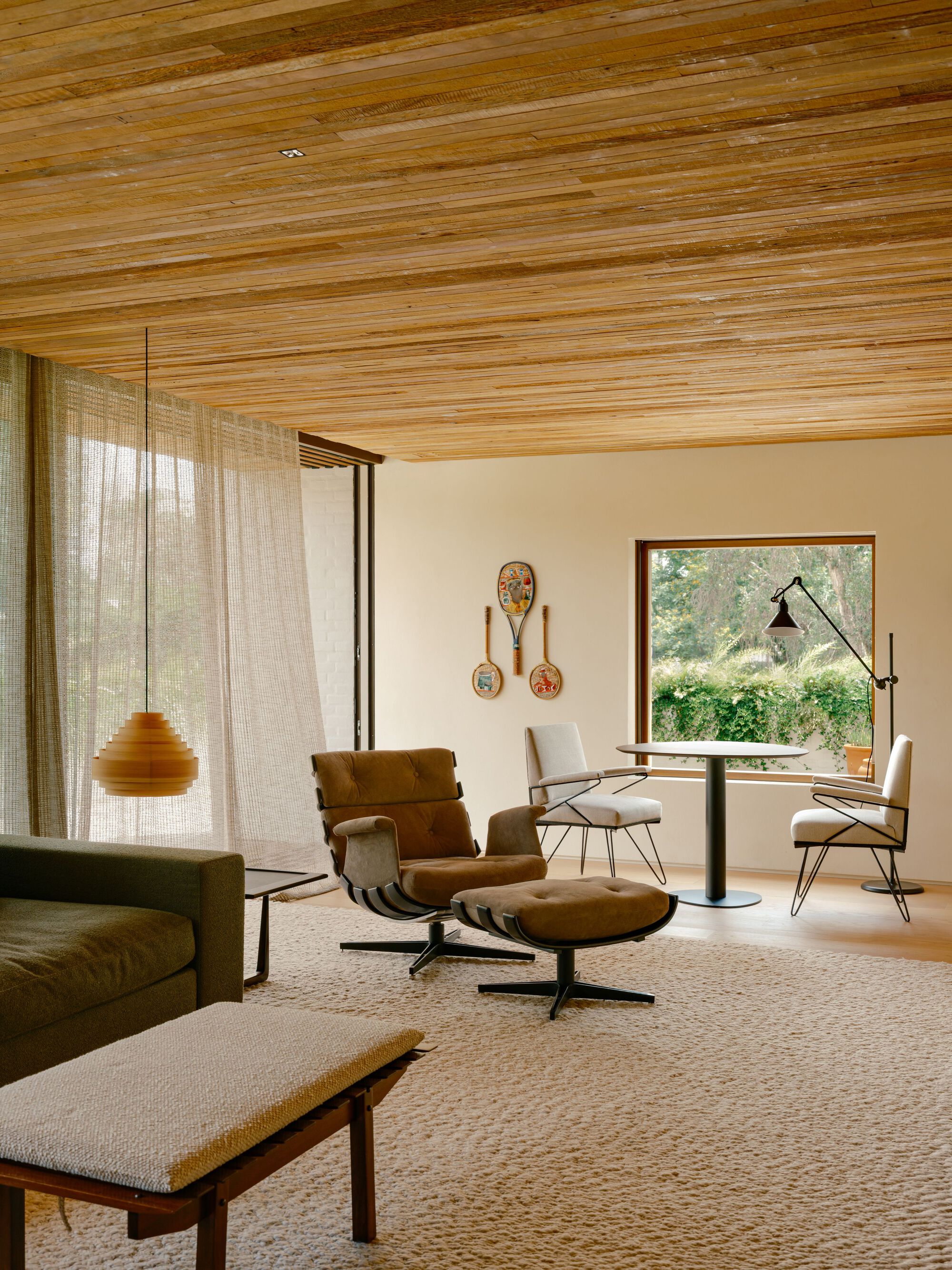 Living area with a reclaimed wood ceiling, leather lounge chair and ottoman, and a square window framing a hedge
