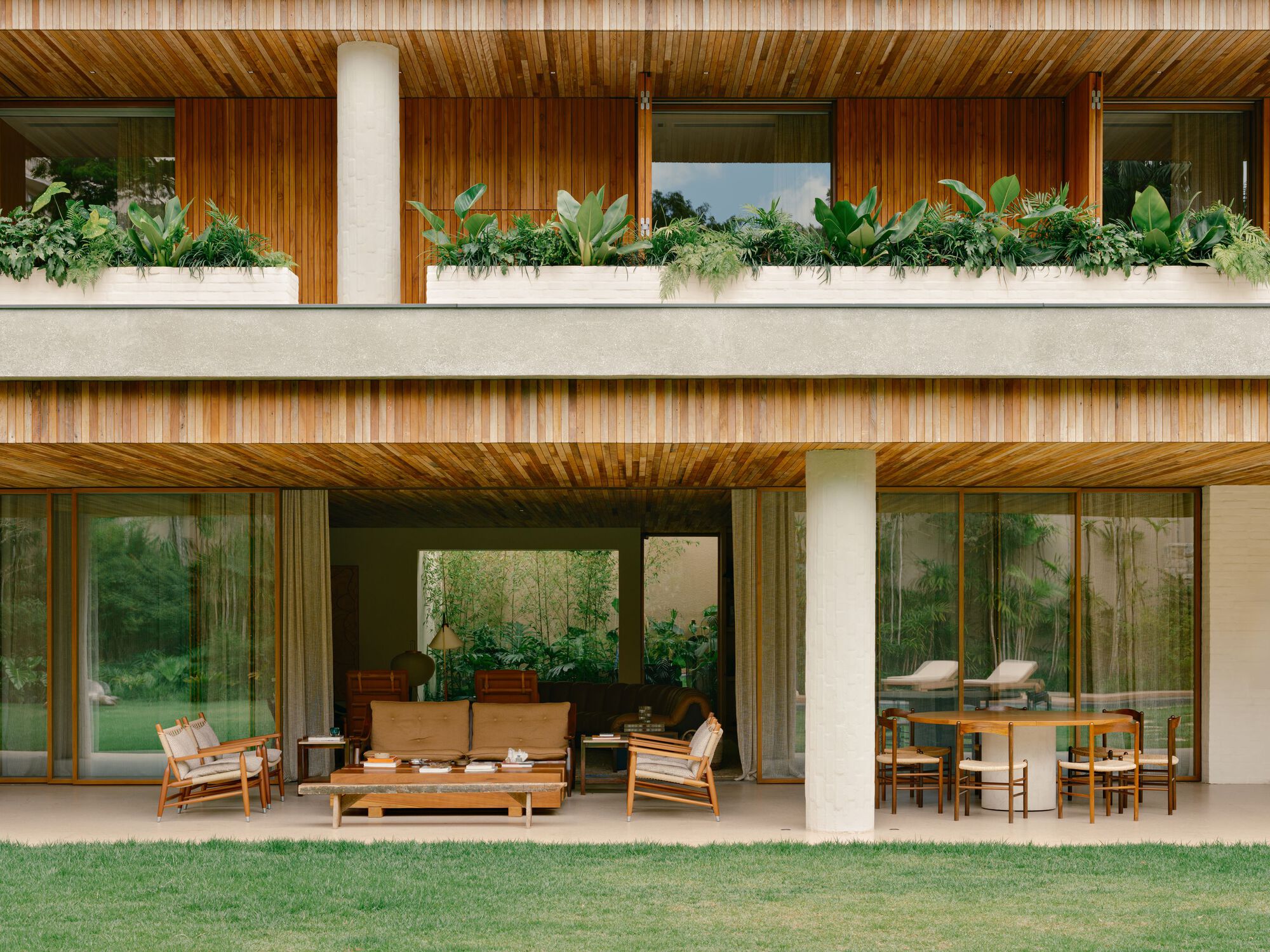 Ground-floor terrace with timber lounge chairs and a round dining table beneath a planter-lined concrete band and slatted wood soffit