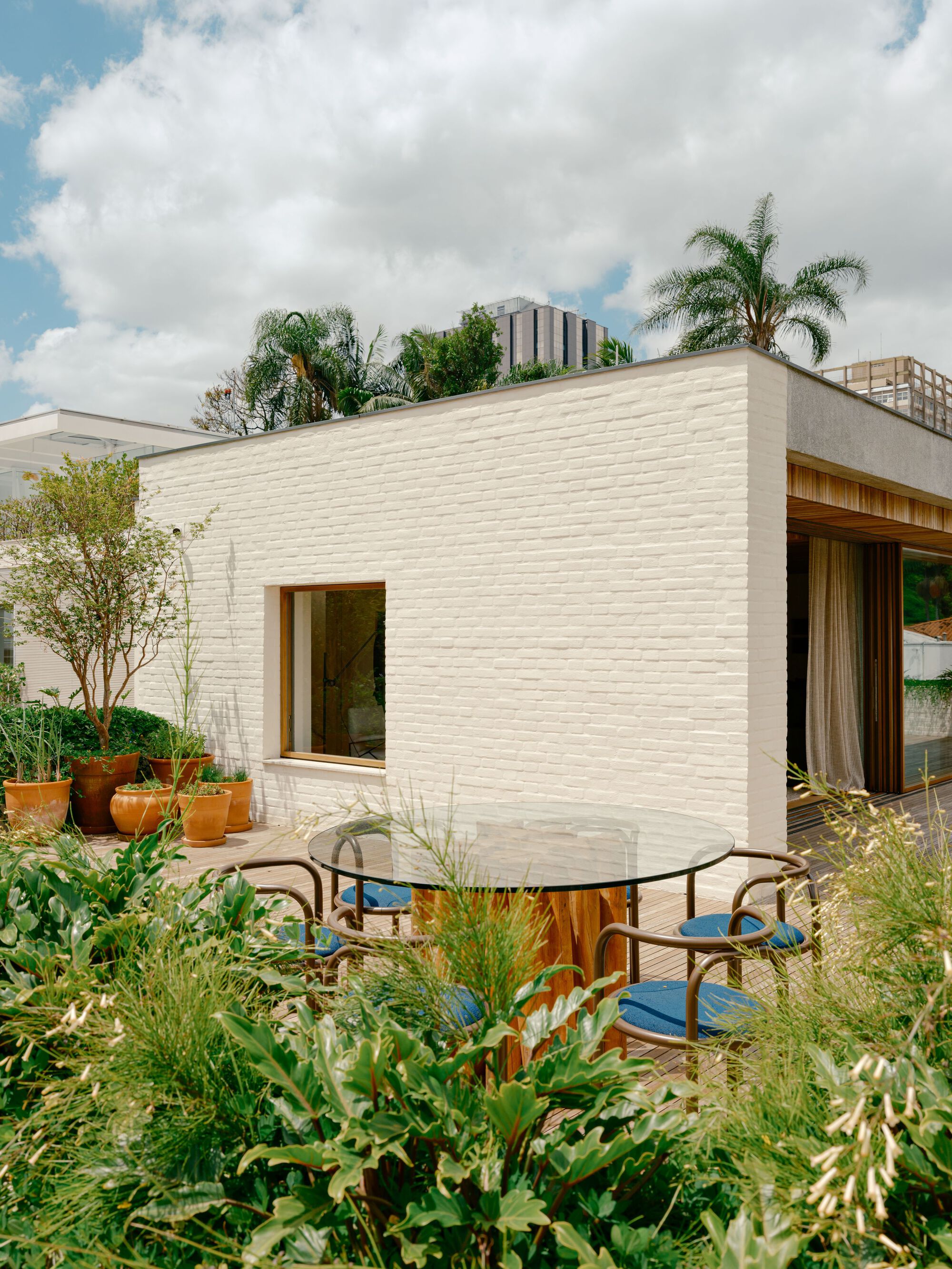 White painted brick volume with a timber-framed window, a glass round table and potted trees on a rooftop terrace under cloudy sky