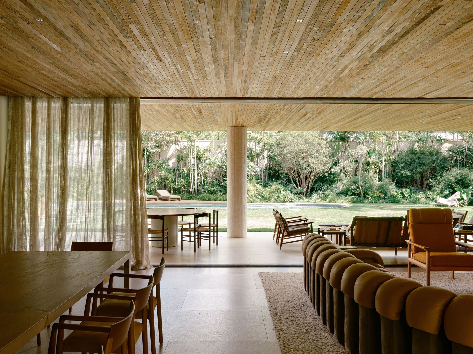 Living area opening through sliding glass to a garden, with a slatted timber ceiling, round column and curved velvet sofa in soft daylight