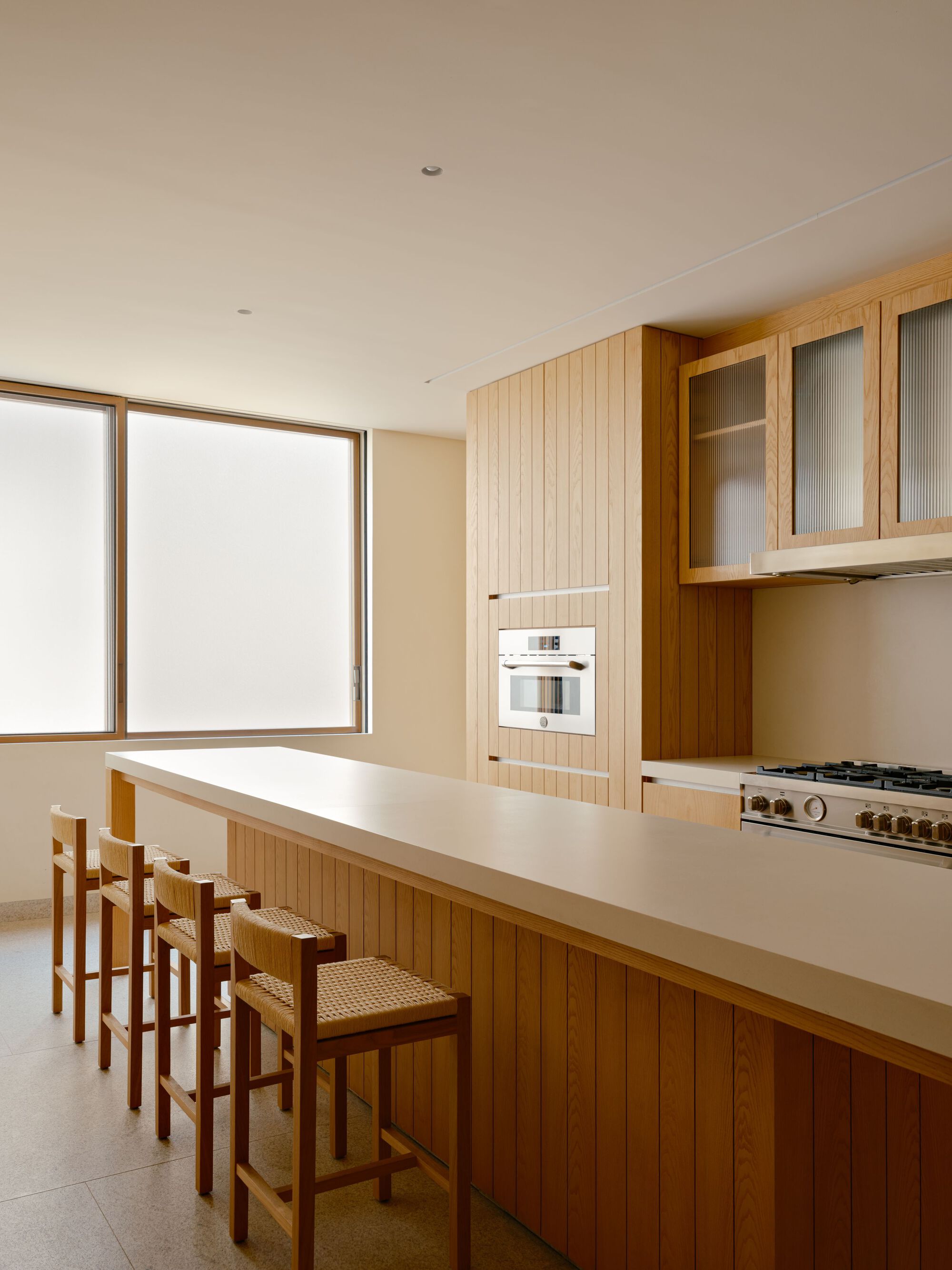 Kitchen with oak-clad island, woven-seat stools, vertical timber cabinetry and a built-in oven beside frosted windows glowing with daylight
