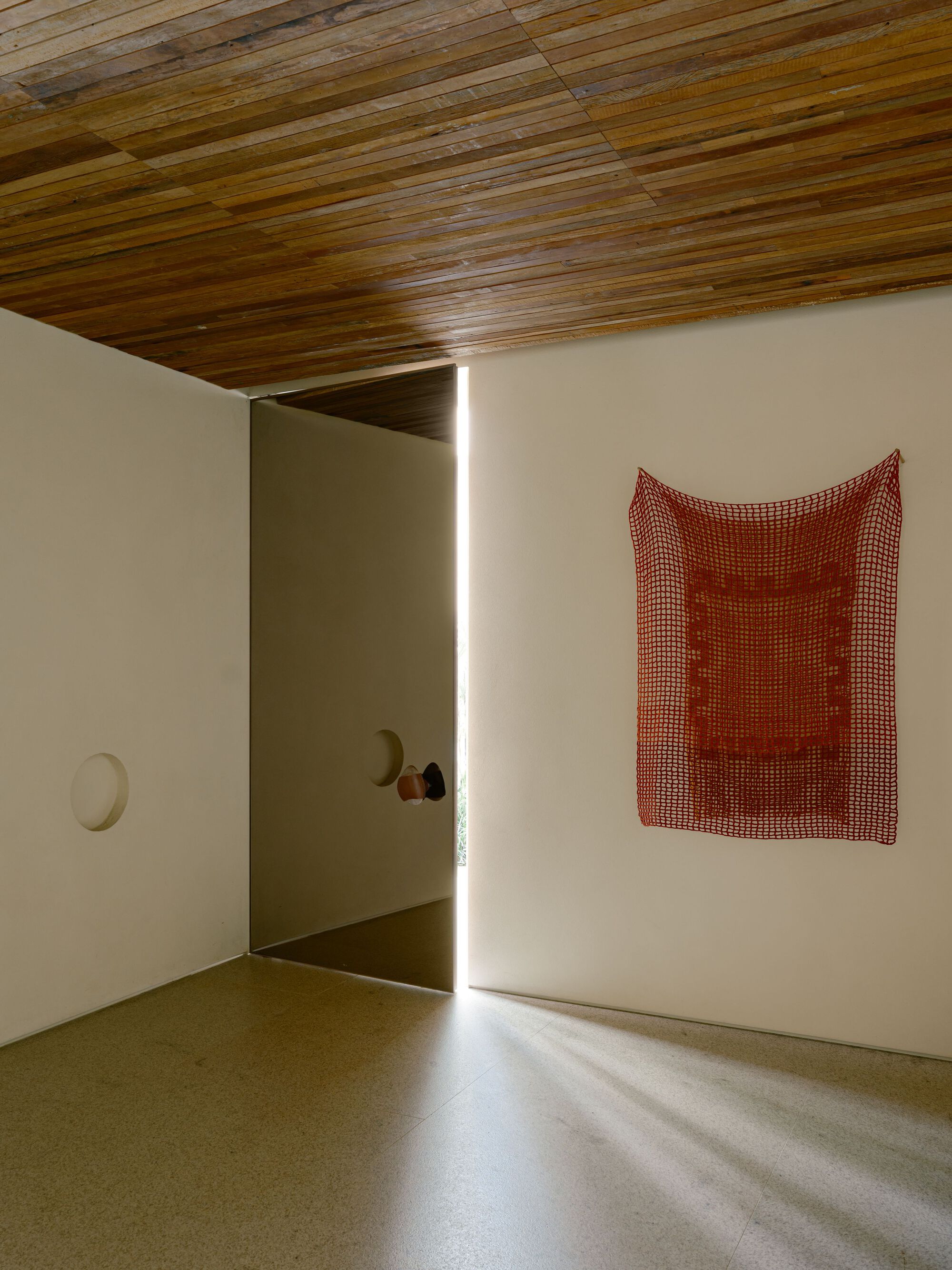 Gallery room with a pivot door cracked open to a sliver of light across the terrazzo floor and a red crocheted hanging on the wall
