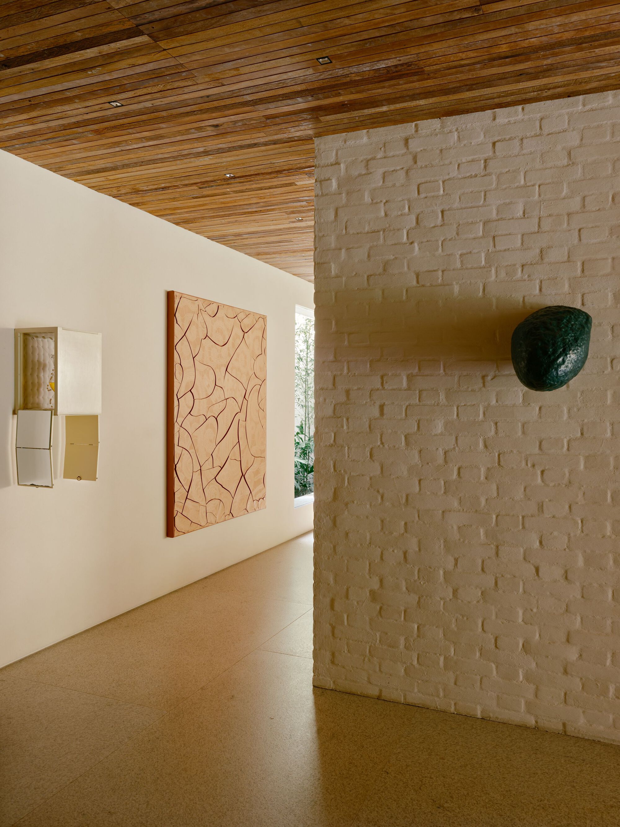 Corridor with a whitewashed brick wall holding a dark green stone sculpture, a carved relief panel opposite and a slatted timber ceiling overhead