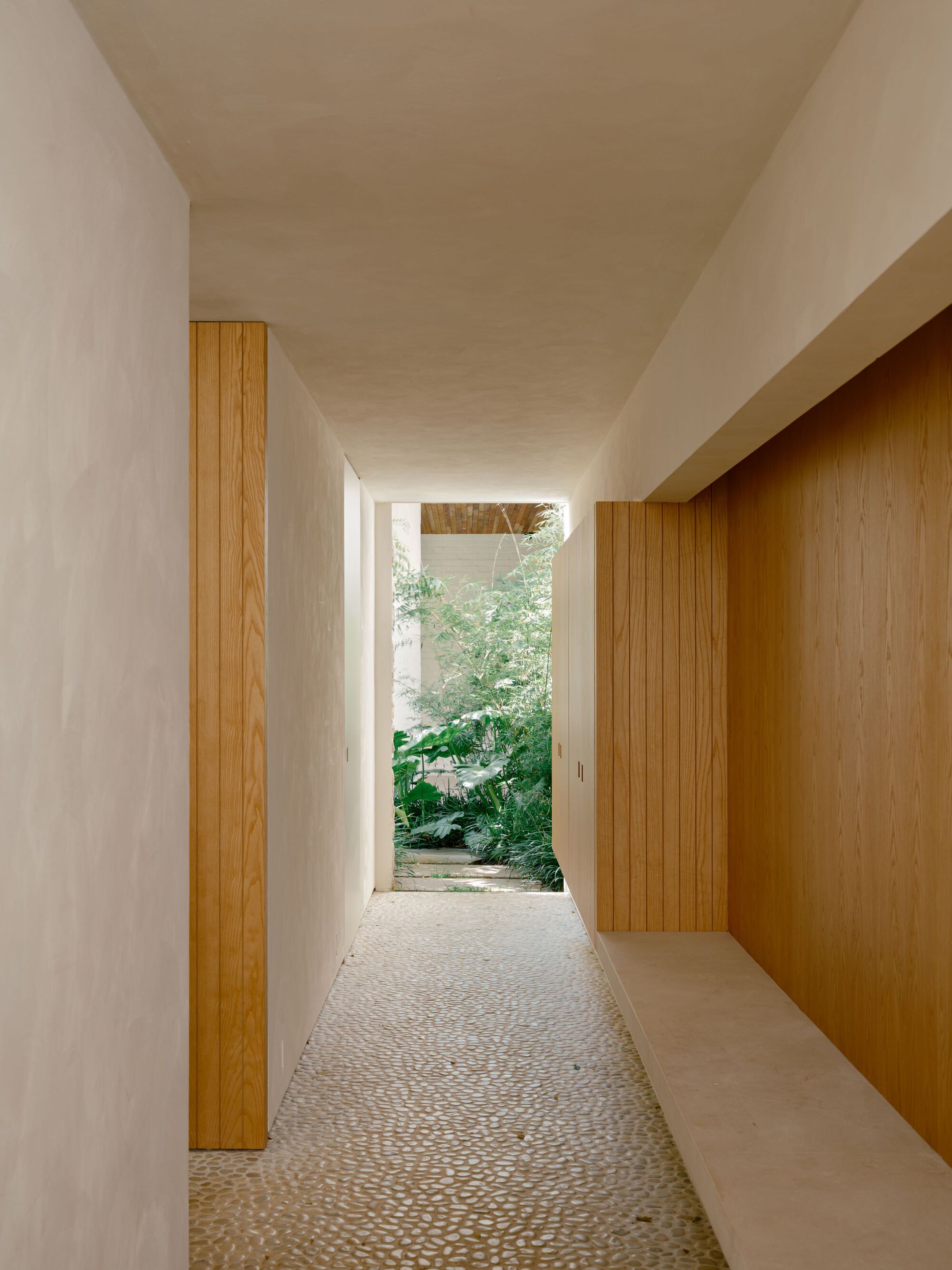 Narrow passage with oak panelling and a built-in stone bench leading over a pebble floor toward a planted courtyard