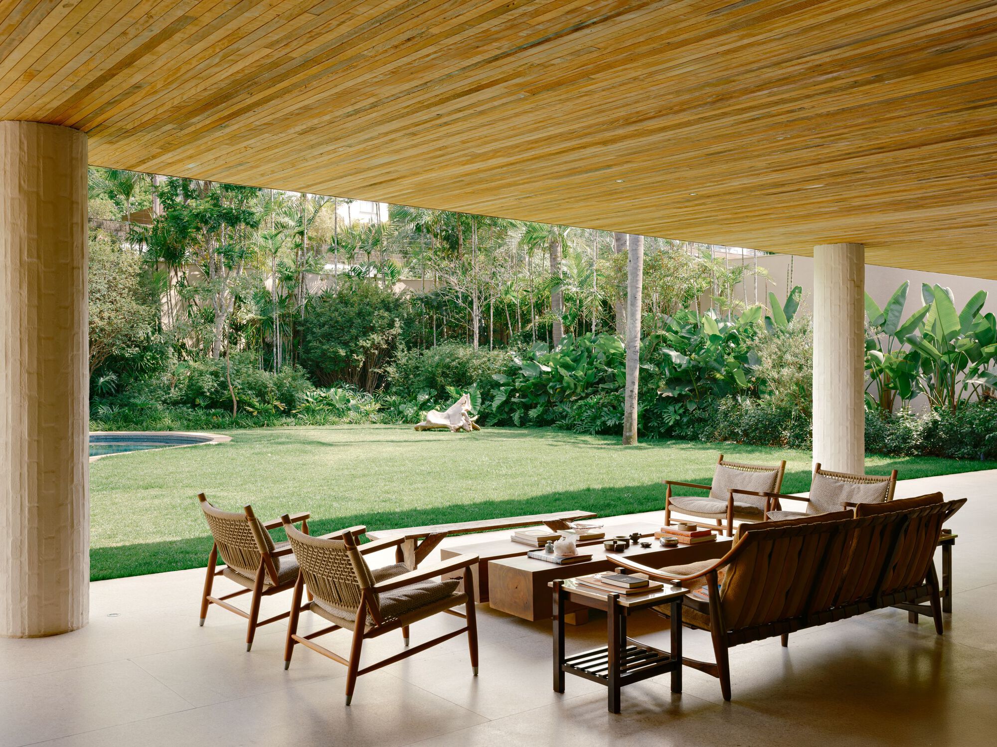 Covered terrace with slatted timber ceiling and round columns, wooden lounge chairs facing a lawn and banana plants in daylight
