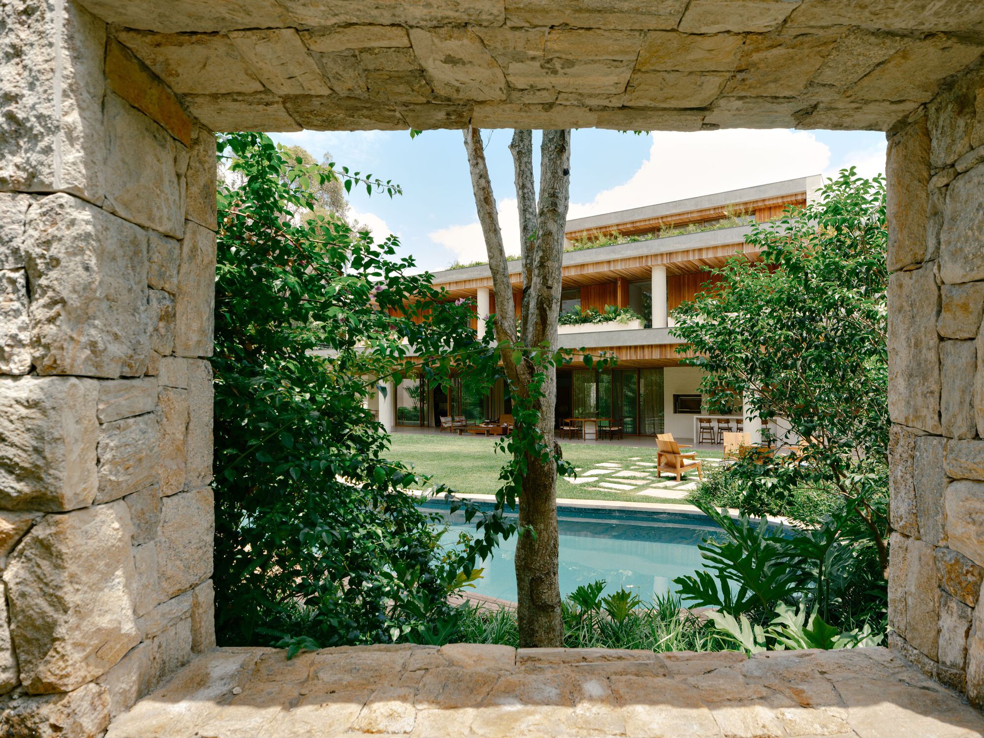Framed view through a rough stone wall opening to a pool, a tree trunk and a timber-clad house with planted terraces