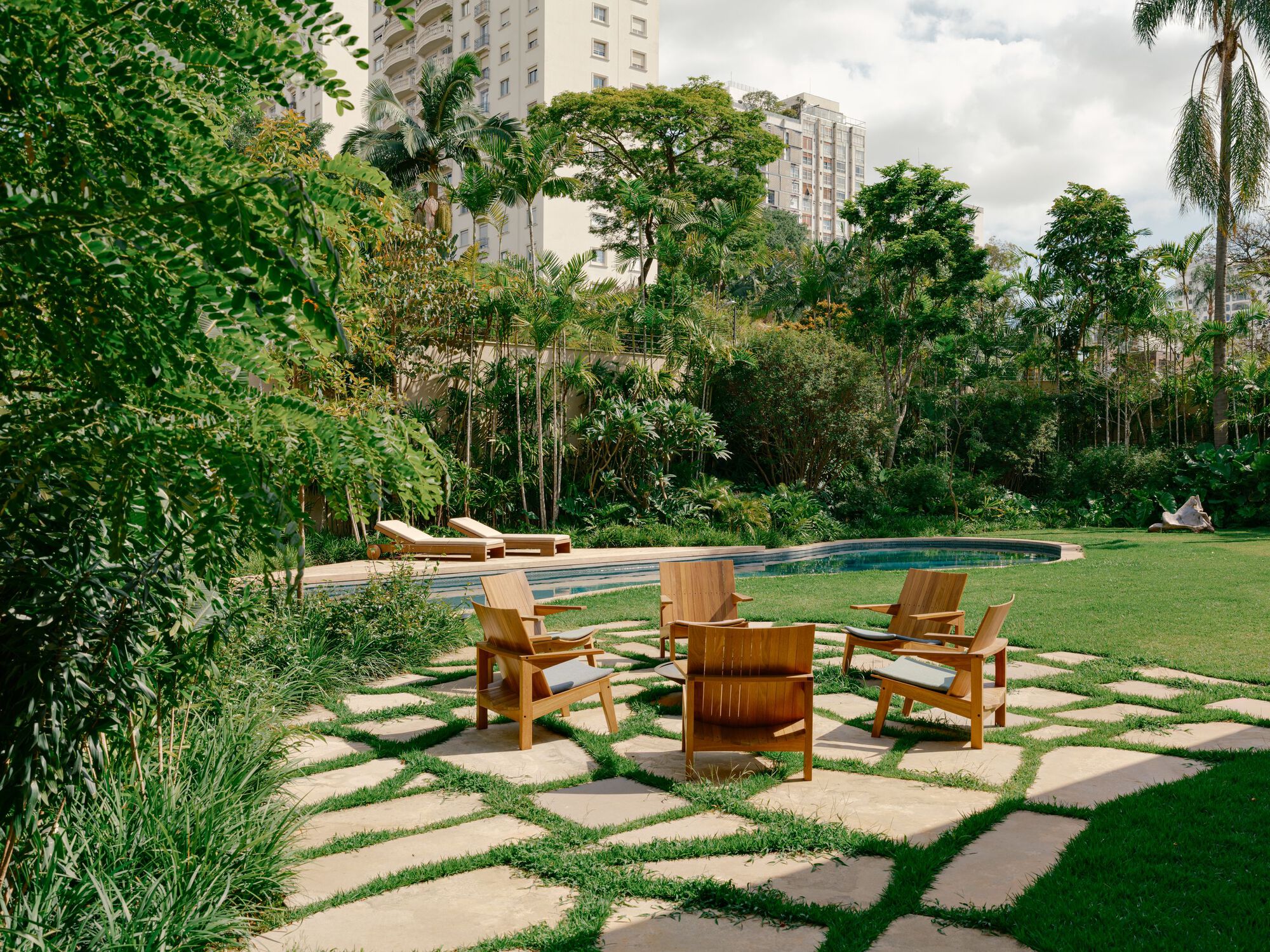 Circle of teak armchairs on irregular stone pavers set in grass, with a curved pool and apartment towers beyond