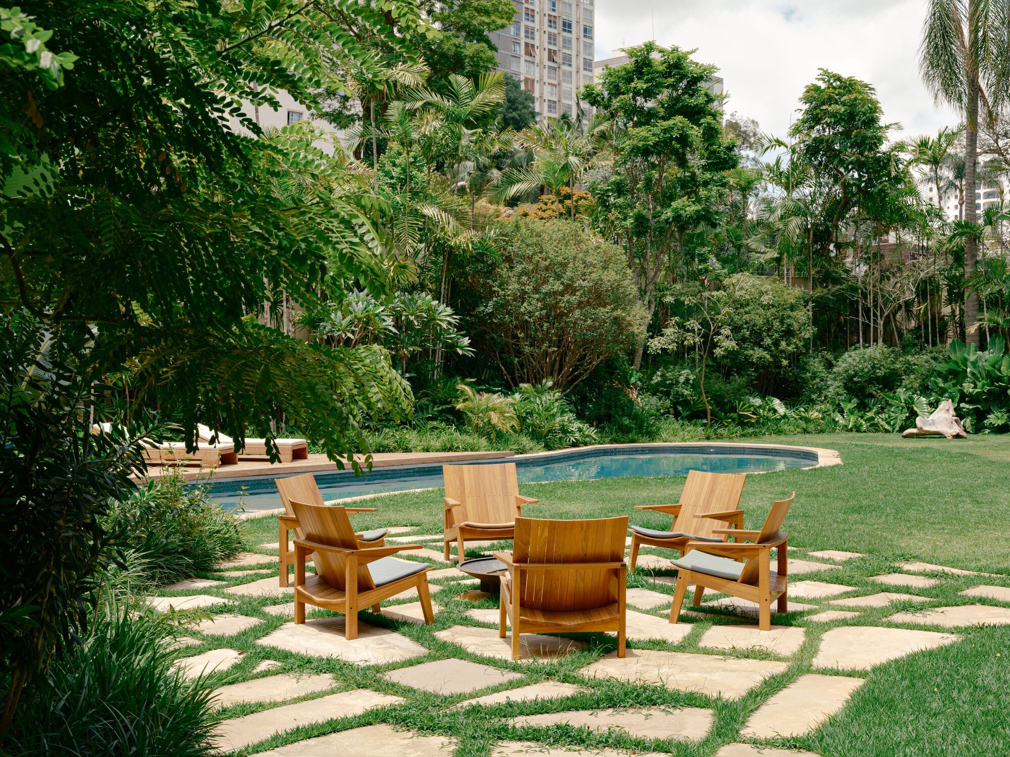 Teak armchairs with grey cushions on stepping stones in lawn, shaded by fern-like foliage with the pool and tall trees behind