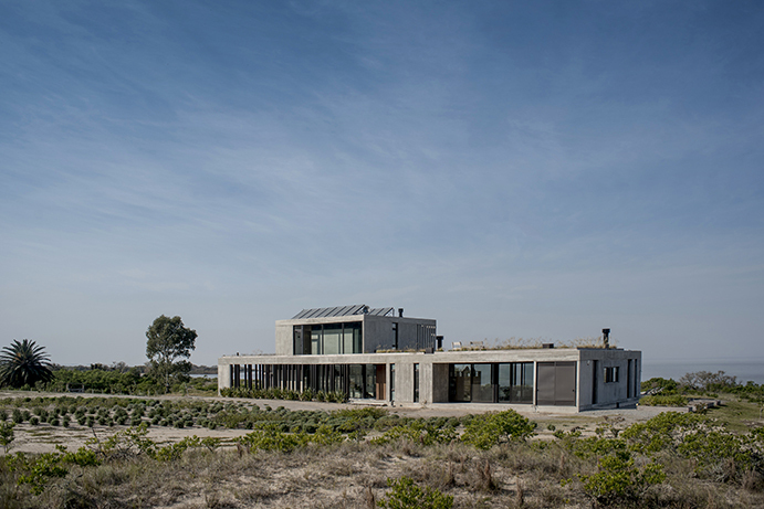 Two-storey board-formed concrete house with a glazed ground floor, set among dune scrub under a wide, streaked blue sky