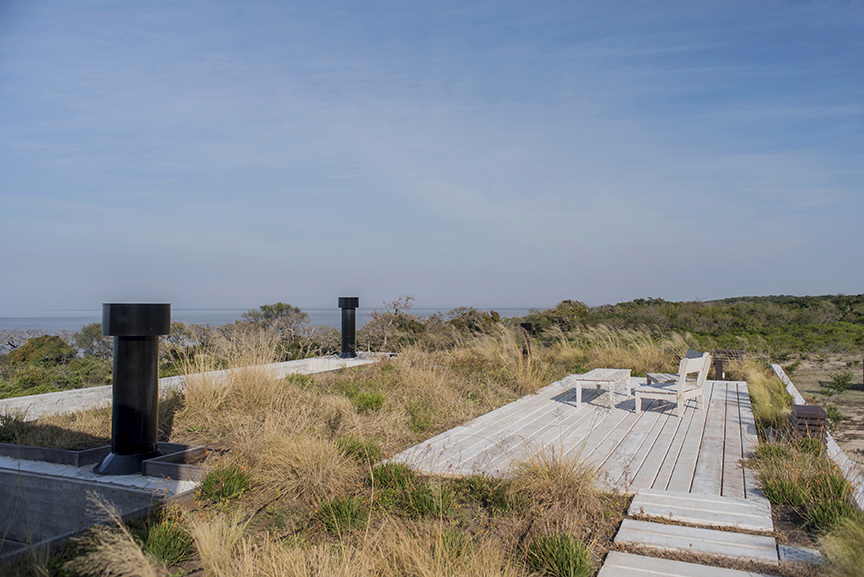 Rooftop timber deck with a bench and low table among tall grasses, black vent stacks and a distant waterline