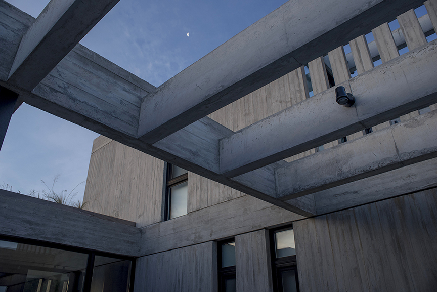 Upward view of concrete pergola beams and board-marked walls with a small crescent moon in the evening sky