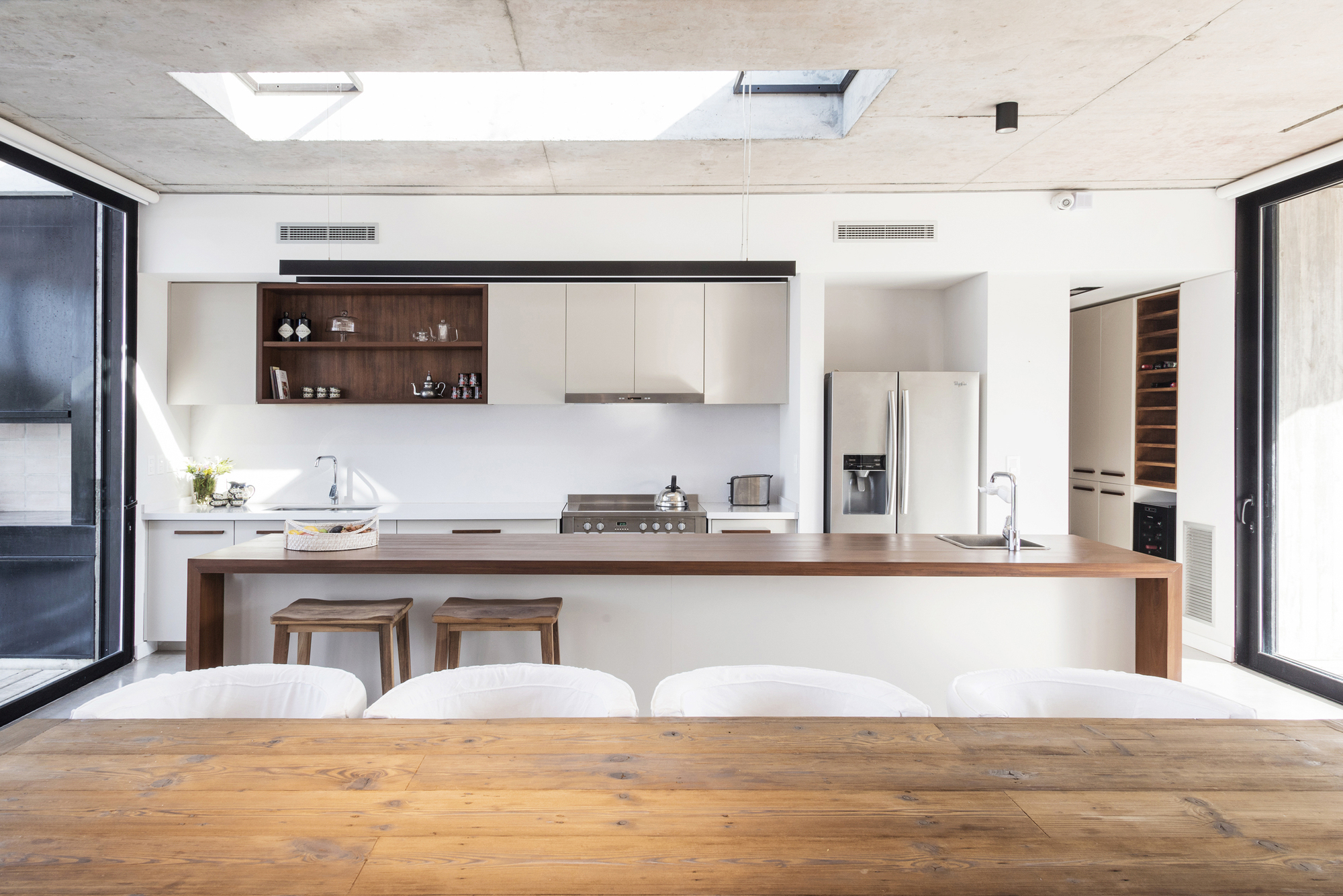 Kitchen with a timber-topped island, white cabinetry and a skylight cut into the concrete ceiling, long plank table in the foreground