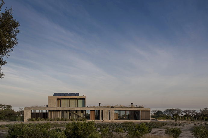 Two-storey board-formed concrete house with rooftop solar panels set among low shrubs under a wide sky of streaked evening cloud