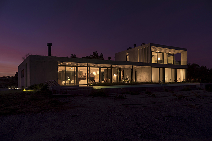 Glazed ground floor of the concrete house glowing from within beneath a purple dusk sky, with a chimney flue above
