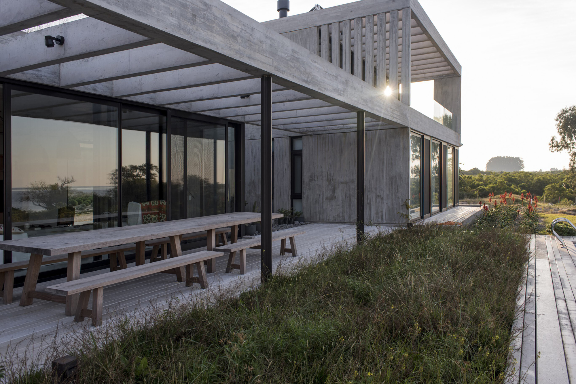 Covered timber deck with a long wooden table and benches beside a concrete pergola and a grassy planted roof edge