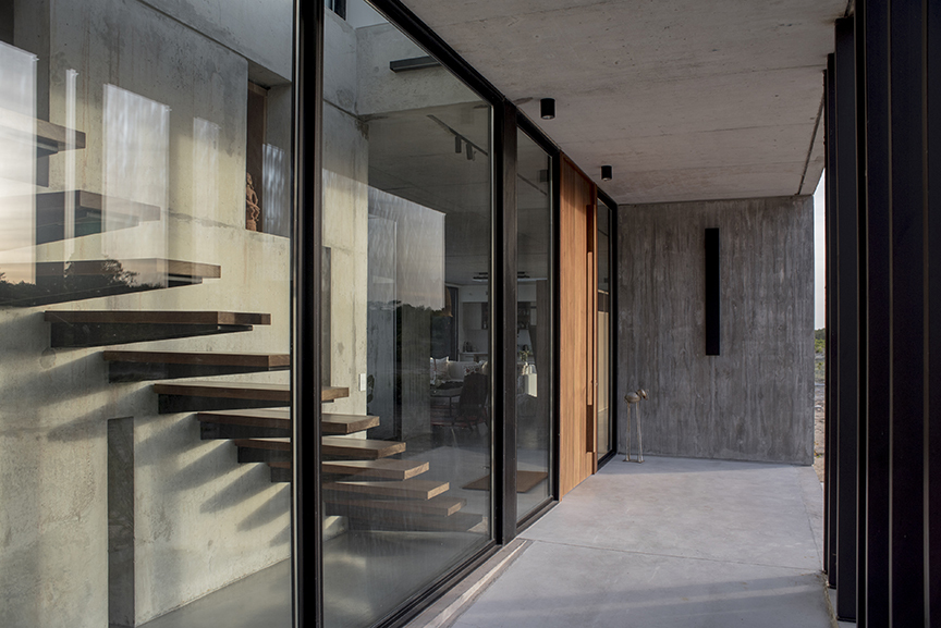 Concrete-floored gallery along a glass wall, with floating timber stair treads reflected in the glazing beside a wood-clad door