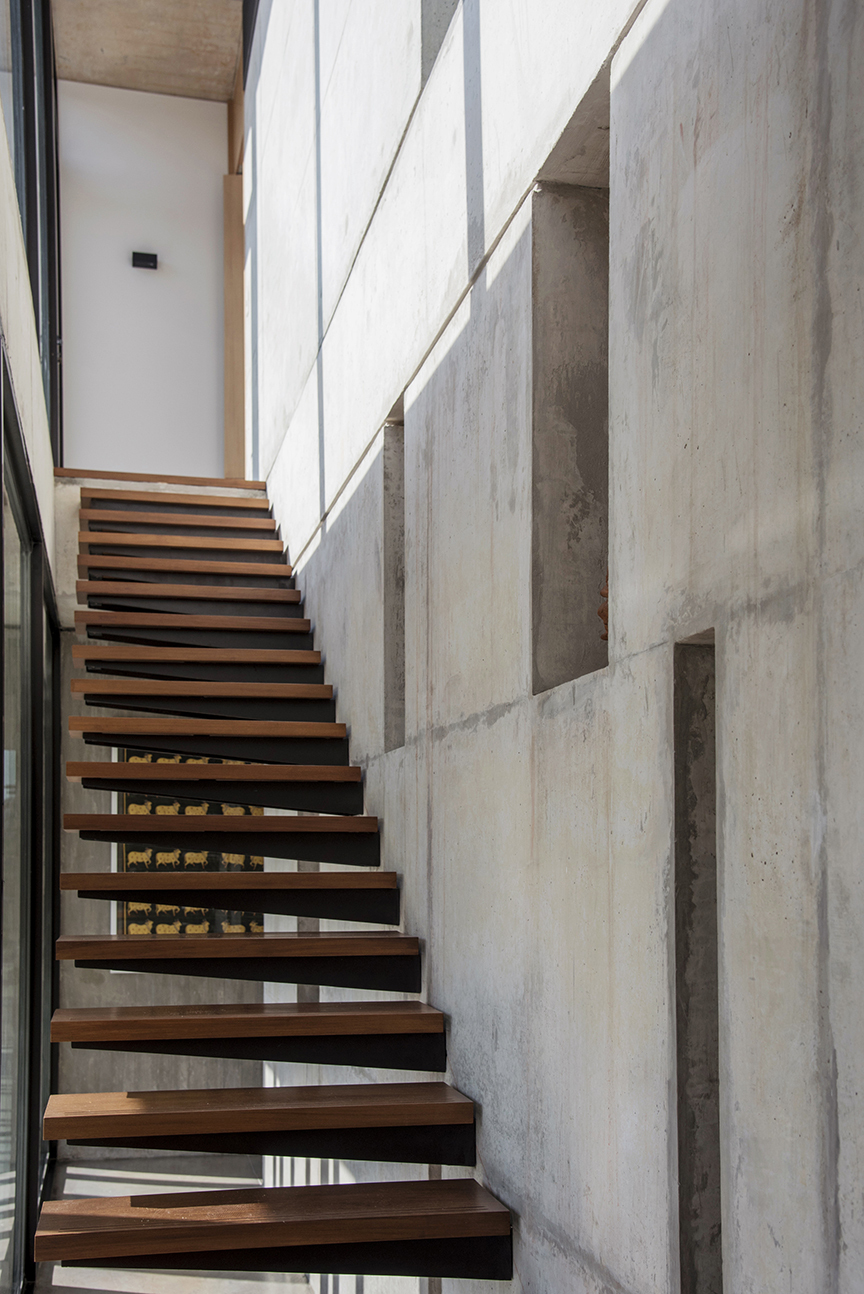 Cantilevered timber stair rising beside a board-marked concrete wall with recessed niches, lit by sunlight from the side