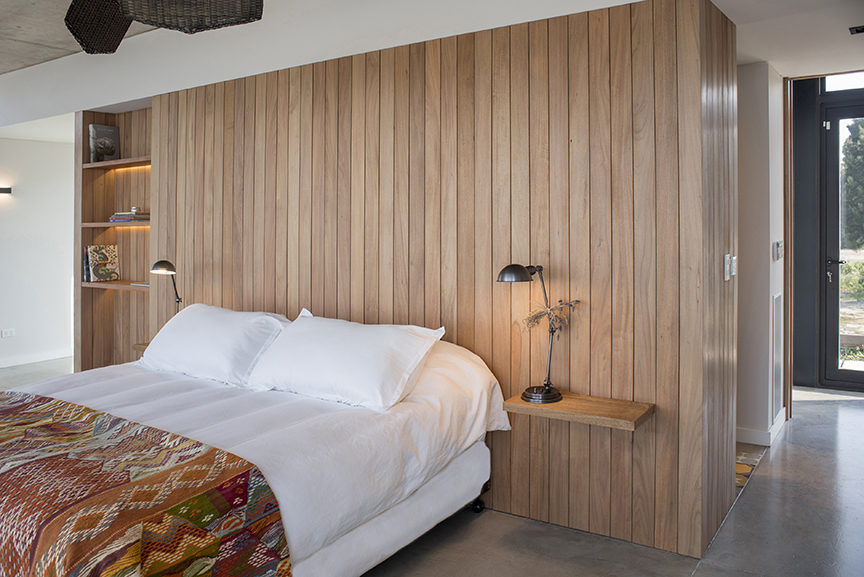Bed against a vertical timber slat partition with a built-in shelf, a small bedside ledge and a metal reading lamp in soft daylight